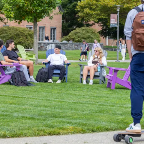 RWU students relaxing in Adirondack chairs on the campus quad, chatting and enjoying a sunny day, while a student on a skateboard passes by in the foreground.
