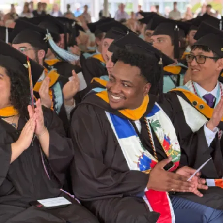 Graduates in caps and gowns seated in rows, smiling and clapping during the 2025 Commencement ceremony at Roger Williams University.