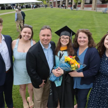 A graduate in cap and gown smiles with a bouquet of flowers, surrounded by family members on the Commons Quad at Roger Williams University during Commencement 2025.