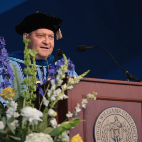 Roger Williams University President Ioannis Miaoulis, dressed in academic regalia, speaks at the podium during Commencement, surrounded by floral arrangements and the university seal.