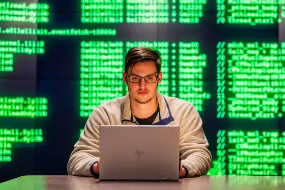 Student sitting at desk, in front of a background of various code, looking at laptop