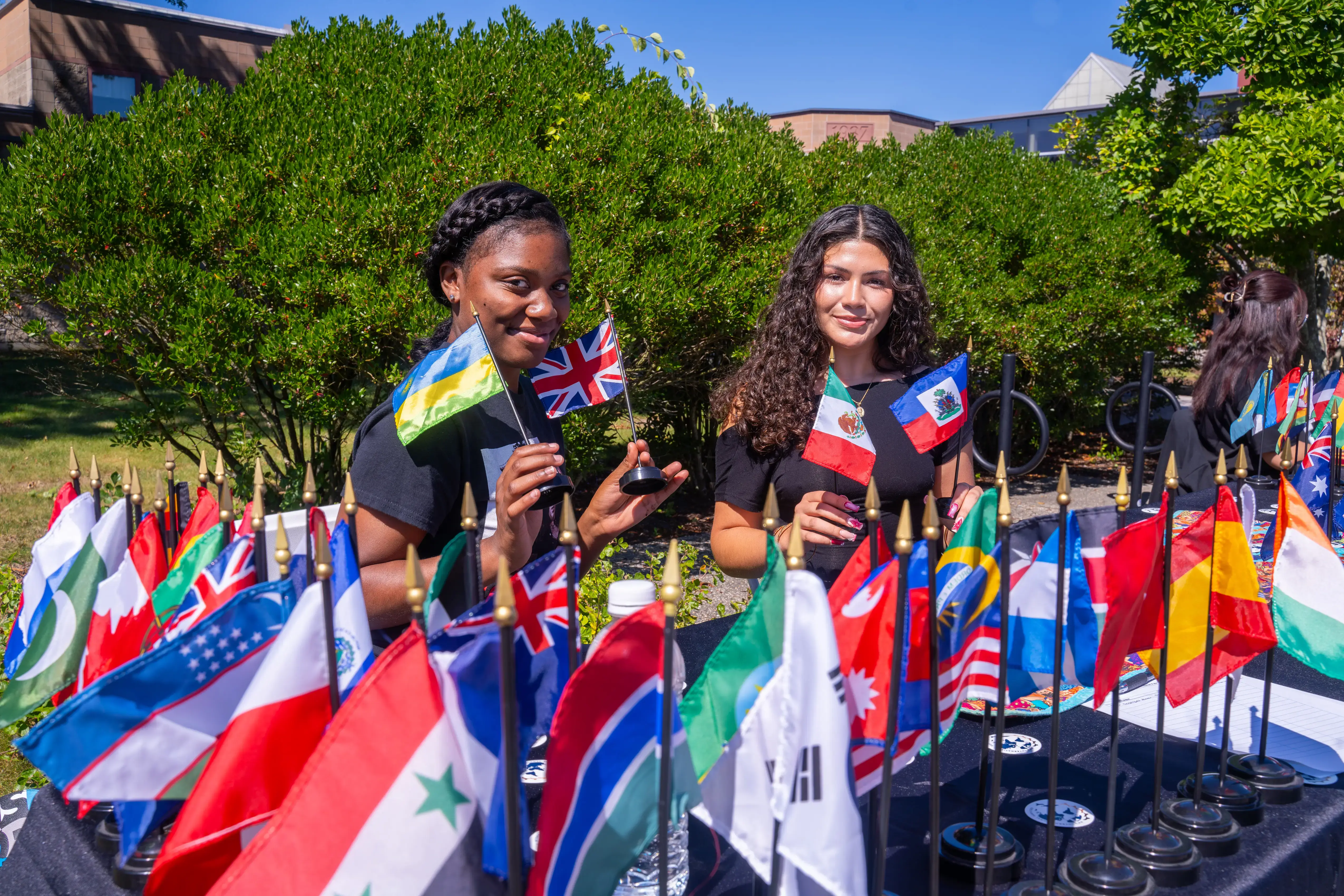 Two women hold mini flags of Ukraine, UK, Mexico, and Haiti at a table with international flags during an outdoor event.