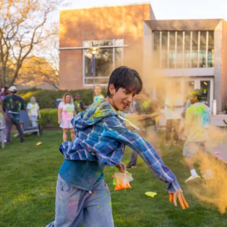 A student throws vibrant orange powder during Roger Williams University’s Holi celebration, surrounded by others covered in colorful powder on the campus lawn in front of The Commons.