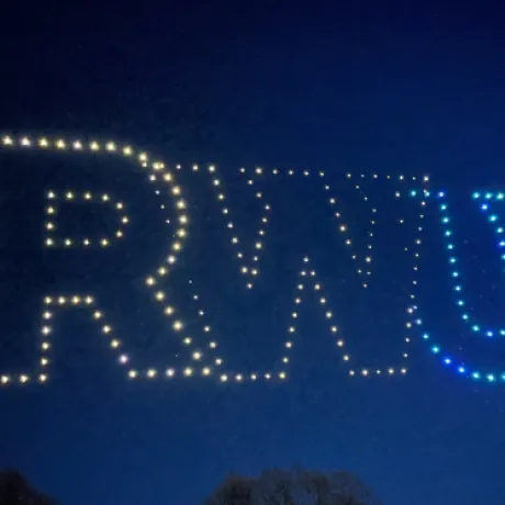 A nighttime drone show displaying the letters 'RWU' illuminated in the sky above Roger Williams University, with glowing white and blue lights forming the university's initials.