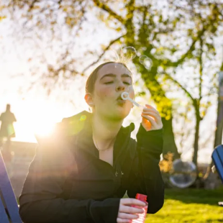 A student blows bubbles while sitting in a blue Adirondack chair on the lawn at Roger Williams University, with the sun setting behind her on the last day of spring classes.