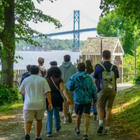 A group of new RWU students walks along a wooded path toward the Mount Hope Bridge during summer orientation. Trees line the trail, and the bridge stretches across the water in the background.
