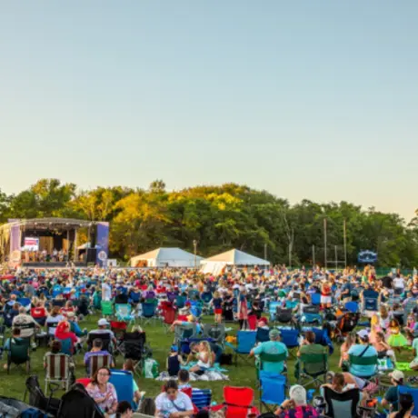 A large crowd sits in lawn chairs on a grassy field at Roger Williams University during the Bristol Fourth of July Concert Series. A stage is set up near the trees, and families and community members are gathered to enjoy the live music.