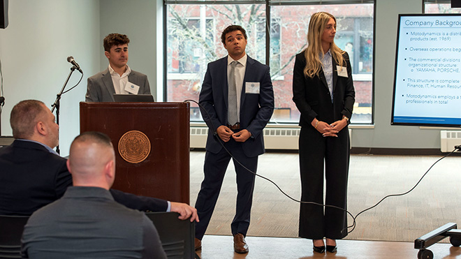 Three MBA students stand and present their findings to an audience