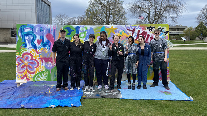 A group of eight people stand in front of the Unity Mural on the quad at RWU