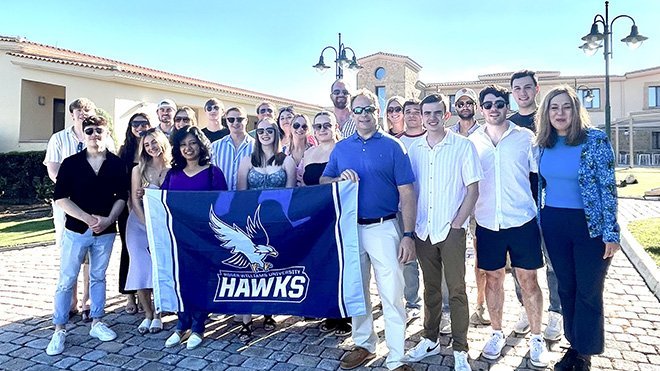 The 2024 MBA cohort with Jay Oliver and Diya Das holding an RWU banner in Greece