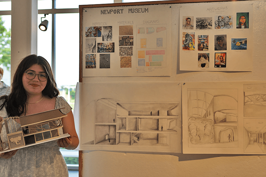 A student standing with her model and her work pinned on a gallery wall 