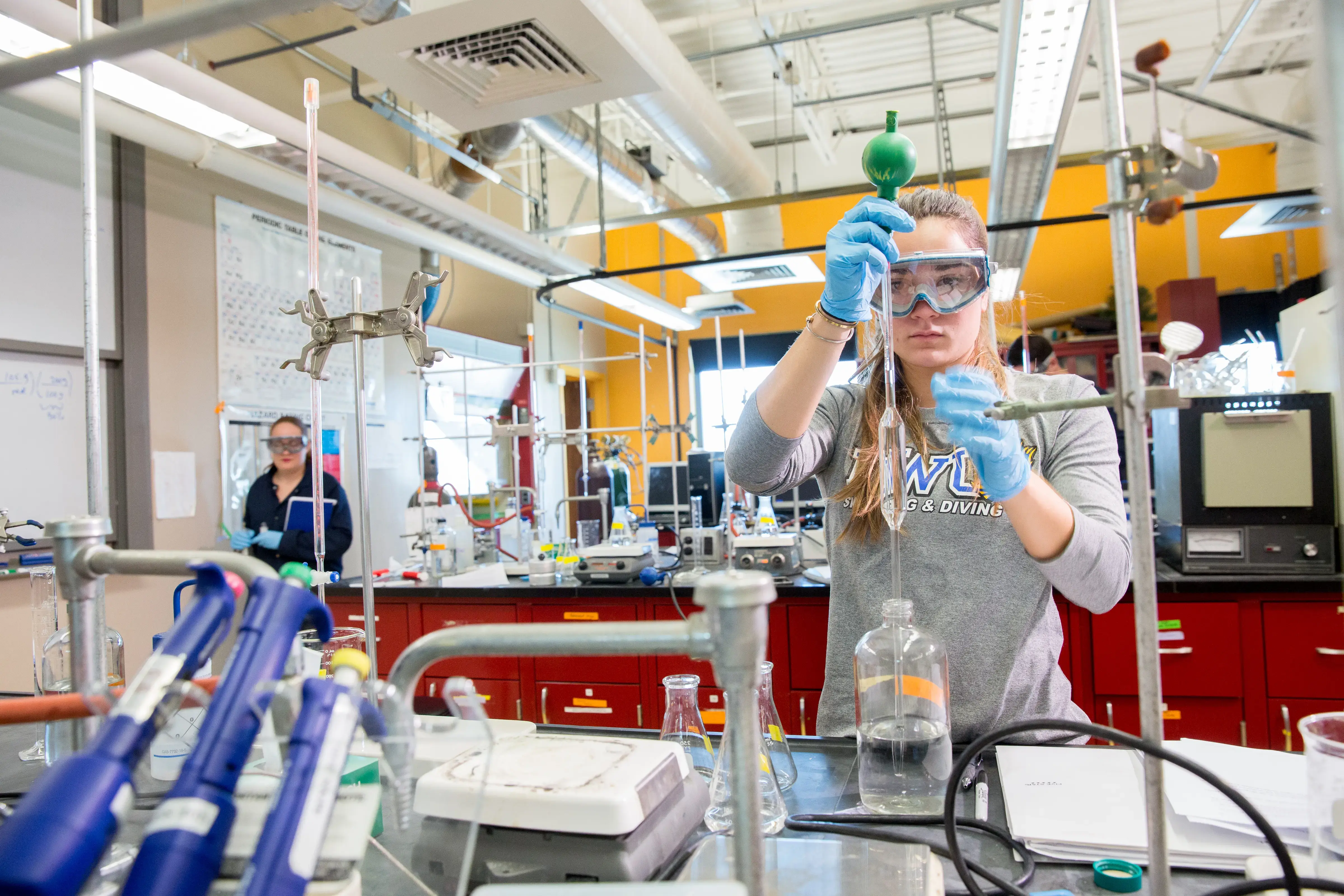 A student in goggles and gloves fills a burette during a chemistry lab, surrounded by flasks and pipettes in a workspace.