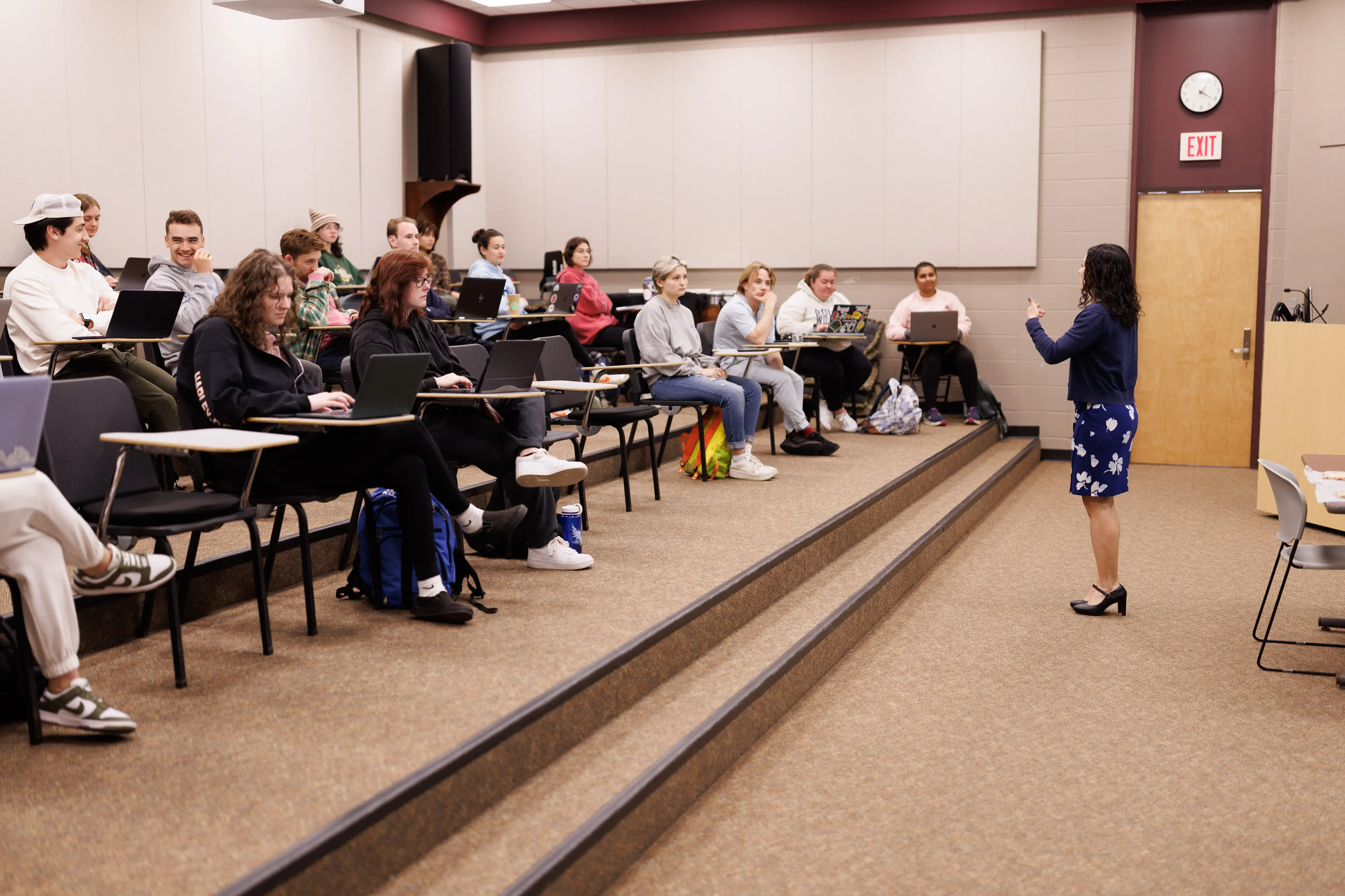 A professor speaks to college students seated with laptops in a tiered lecture hall with beige walls and carpeted floors.