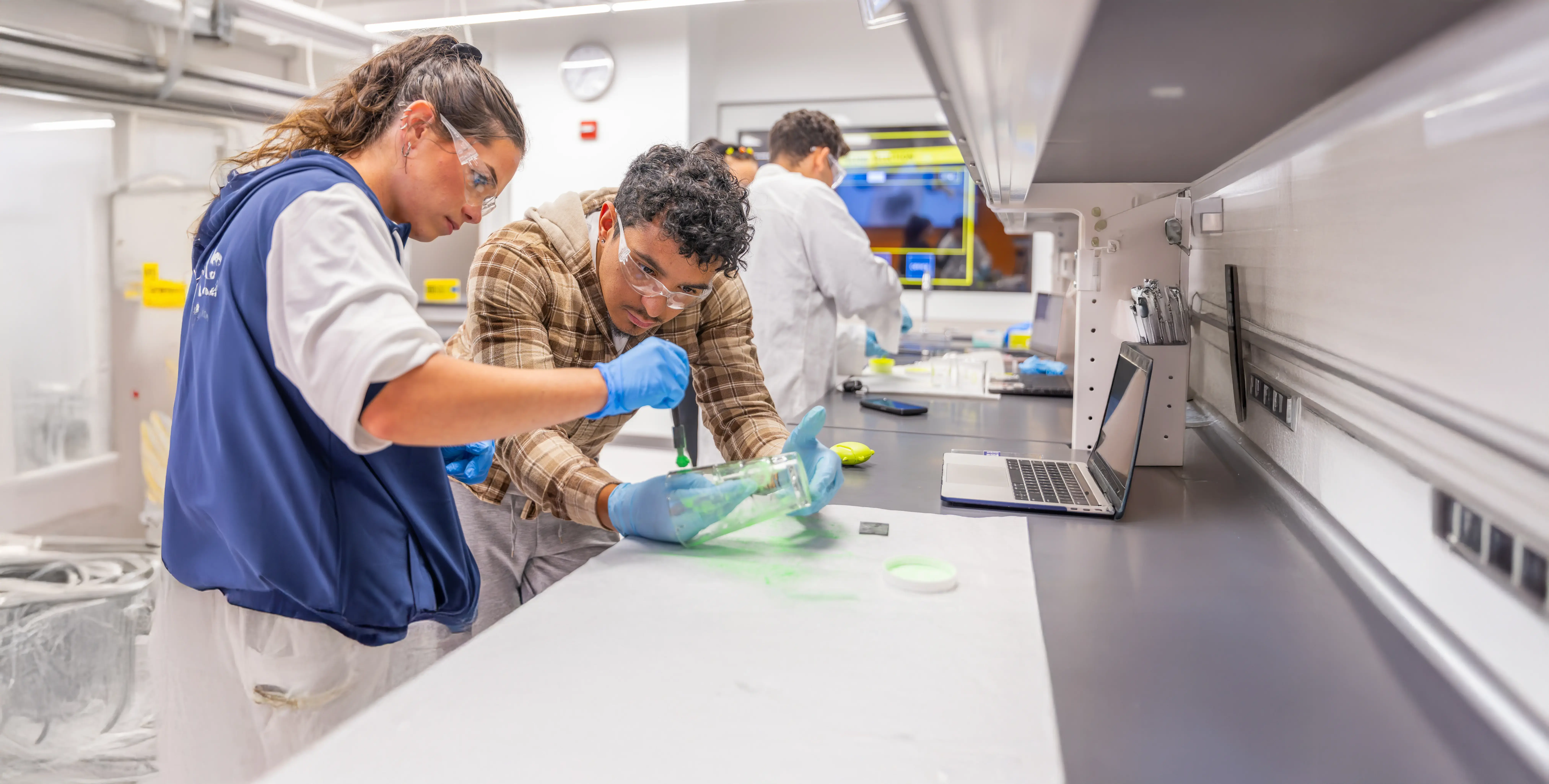 Students in safety gear work with green material in a lab, using tools and laptops at a long workstation.