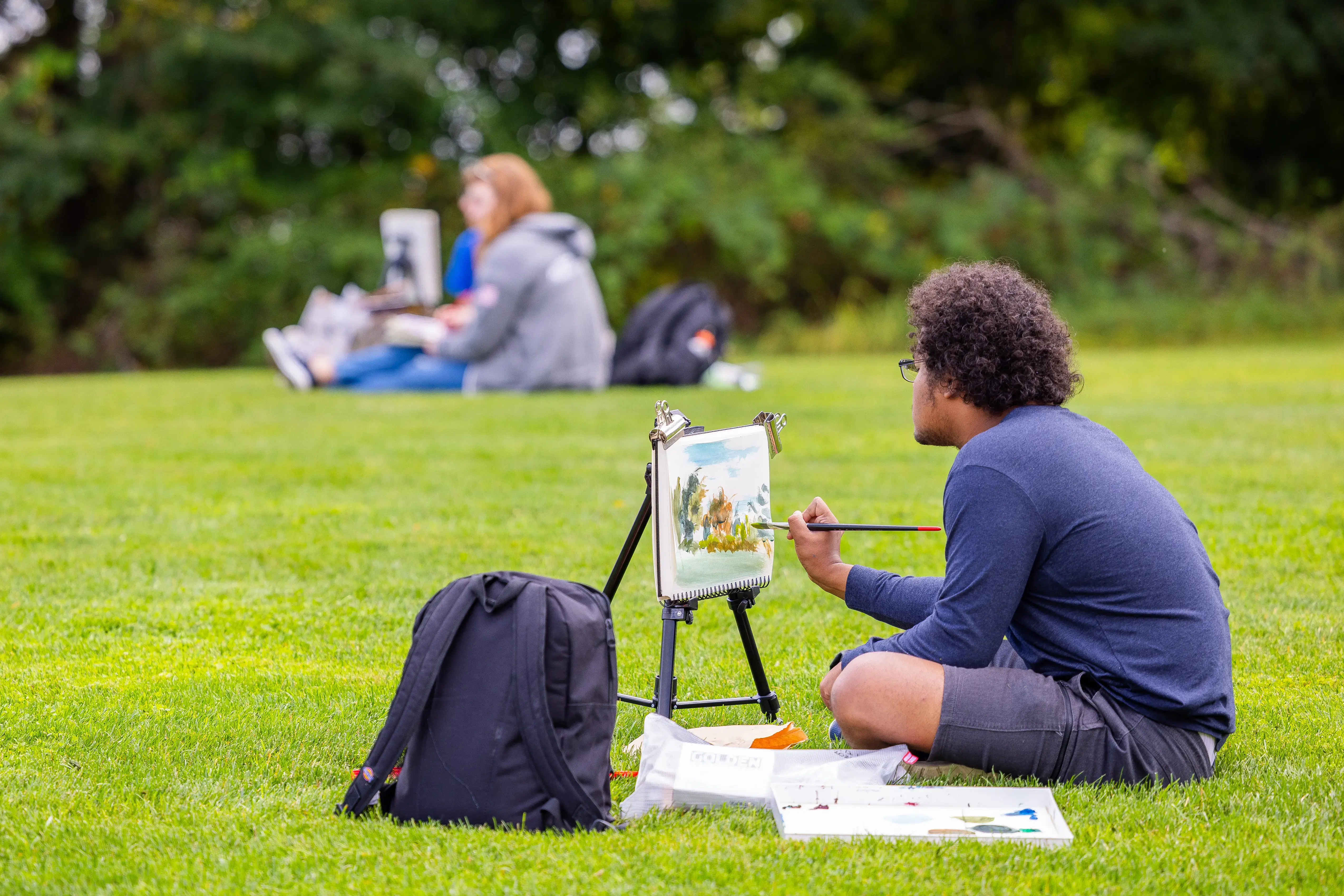 A student paints on a canvas in the grass during an outdoor art class, with supplies and others painting nearby.
