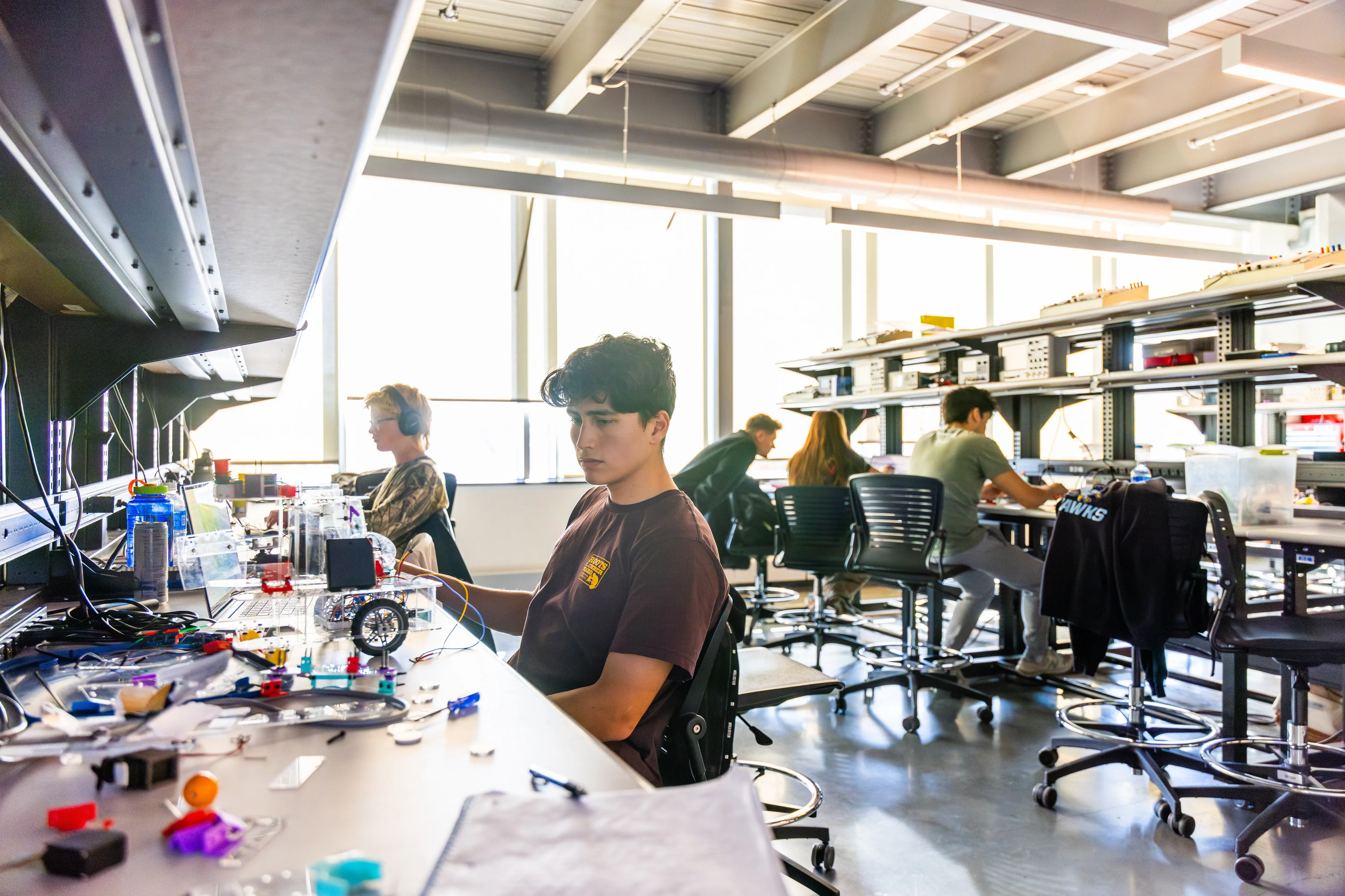 Students work on robotics projects at lab tables, with tools, wires, and parts spread out in a bright, modern classroom.