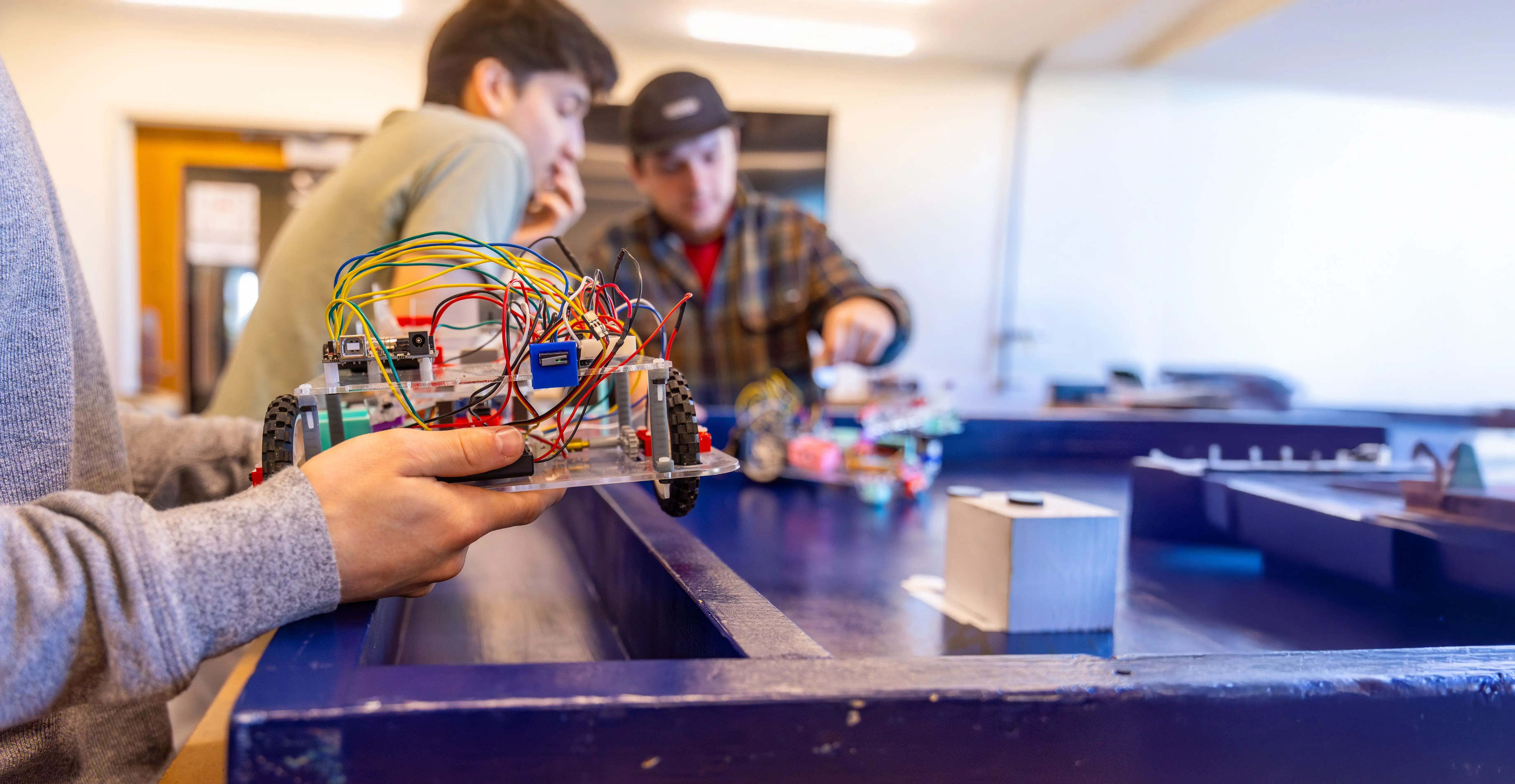 Students work on a robotics project, holding a wheeled device with colorful wires in a hands-on classroom or lab environment.