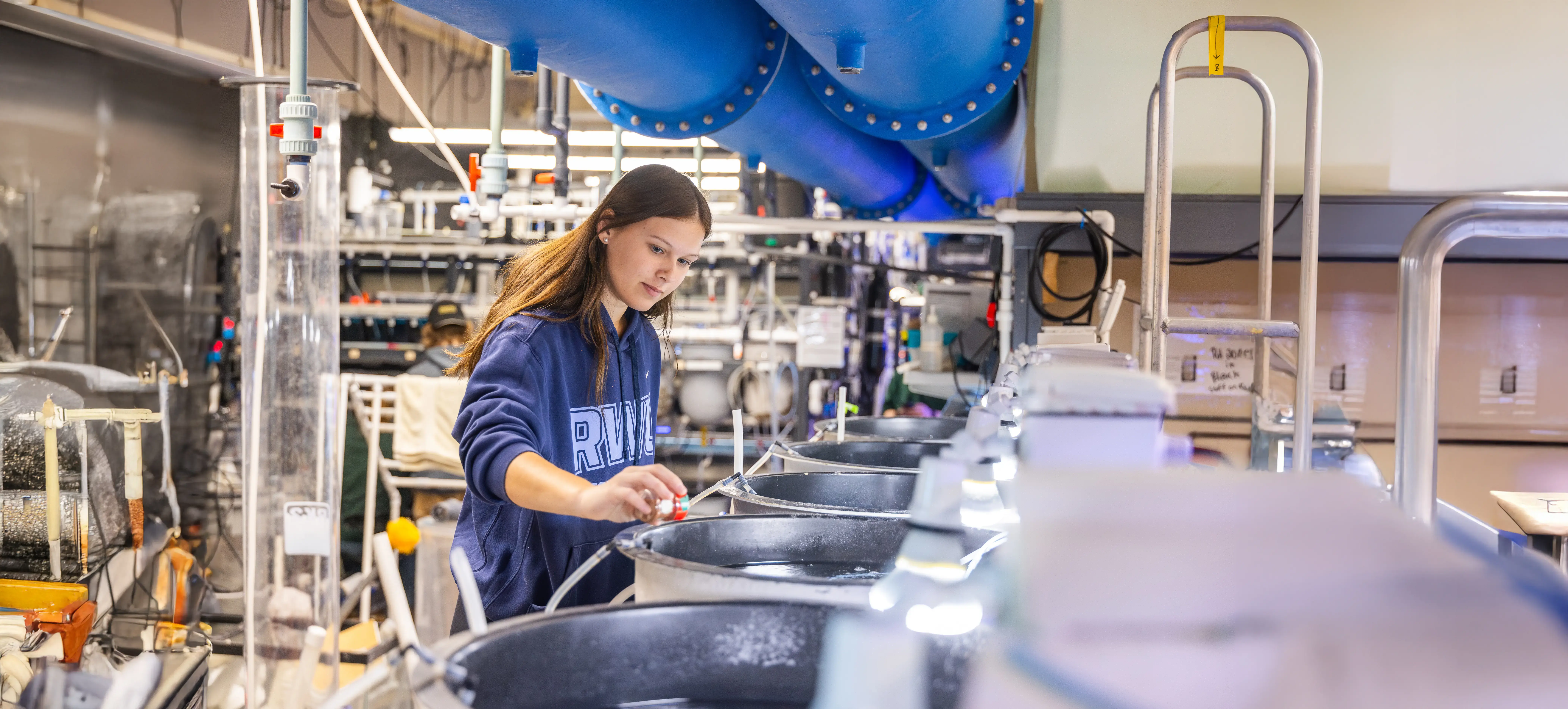A student in the wet lab adjusts equipment near large water tanks and pipes, surrounded by technical instruments.