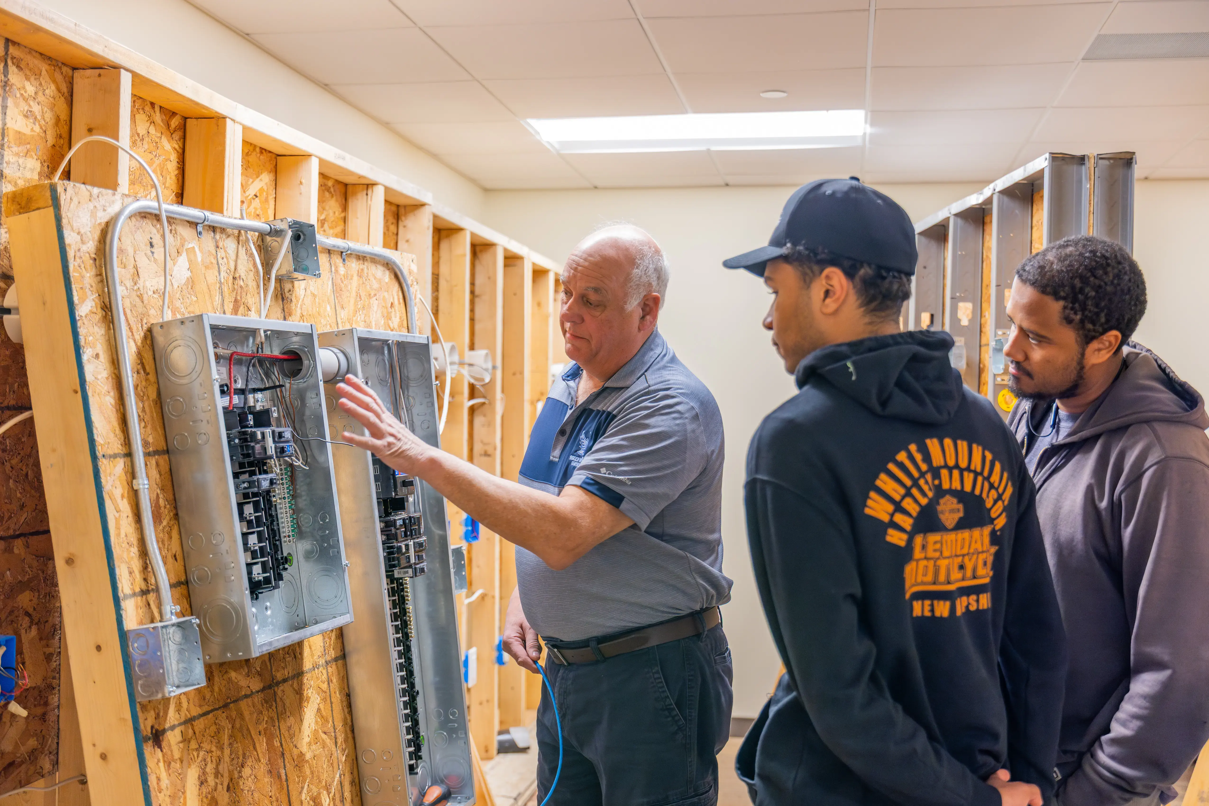 An instructor explains wiring to two students in a workshop with exposed framing, circuit breakers, and electrical panels.