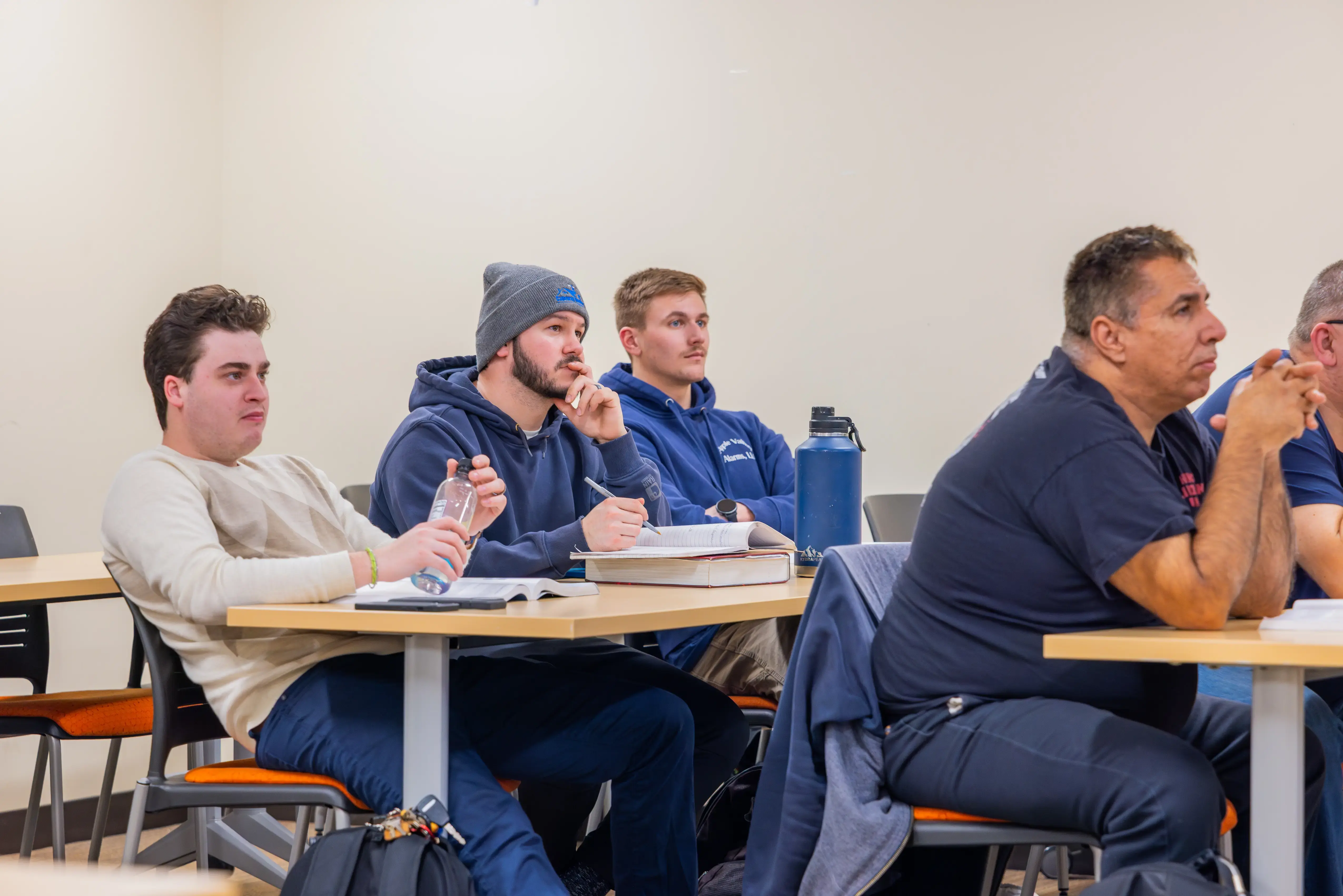 Adult students listen in a vocational classroom, some with notebooks and water bottles, featuring beige walls and chairs.