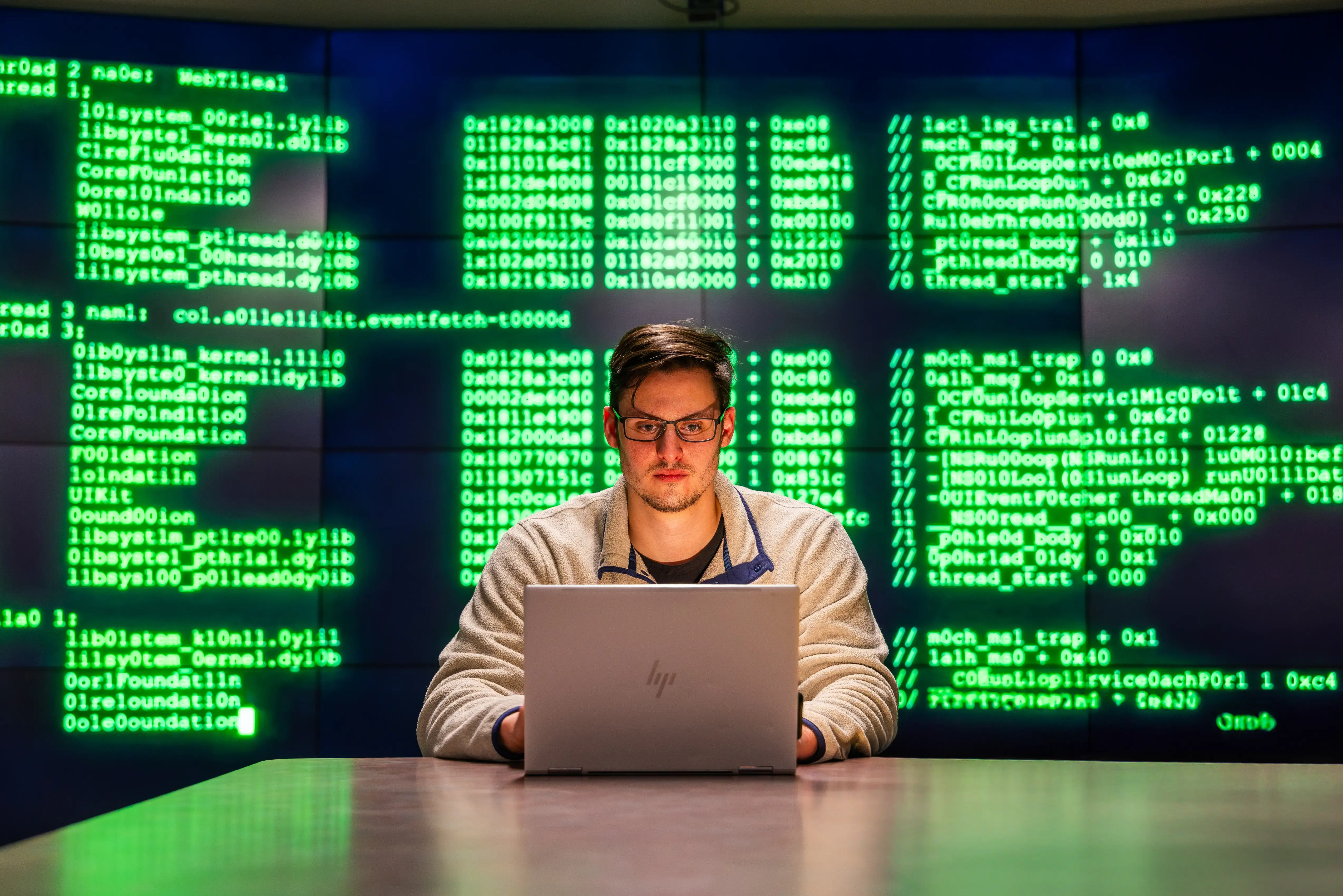 A young man with glasses works on an HP laptop in a dim room with green code displayed on a large screen behind him.