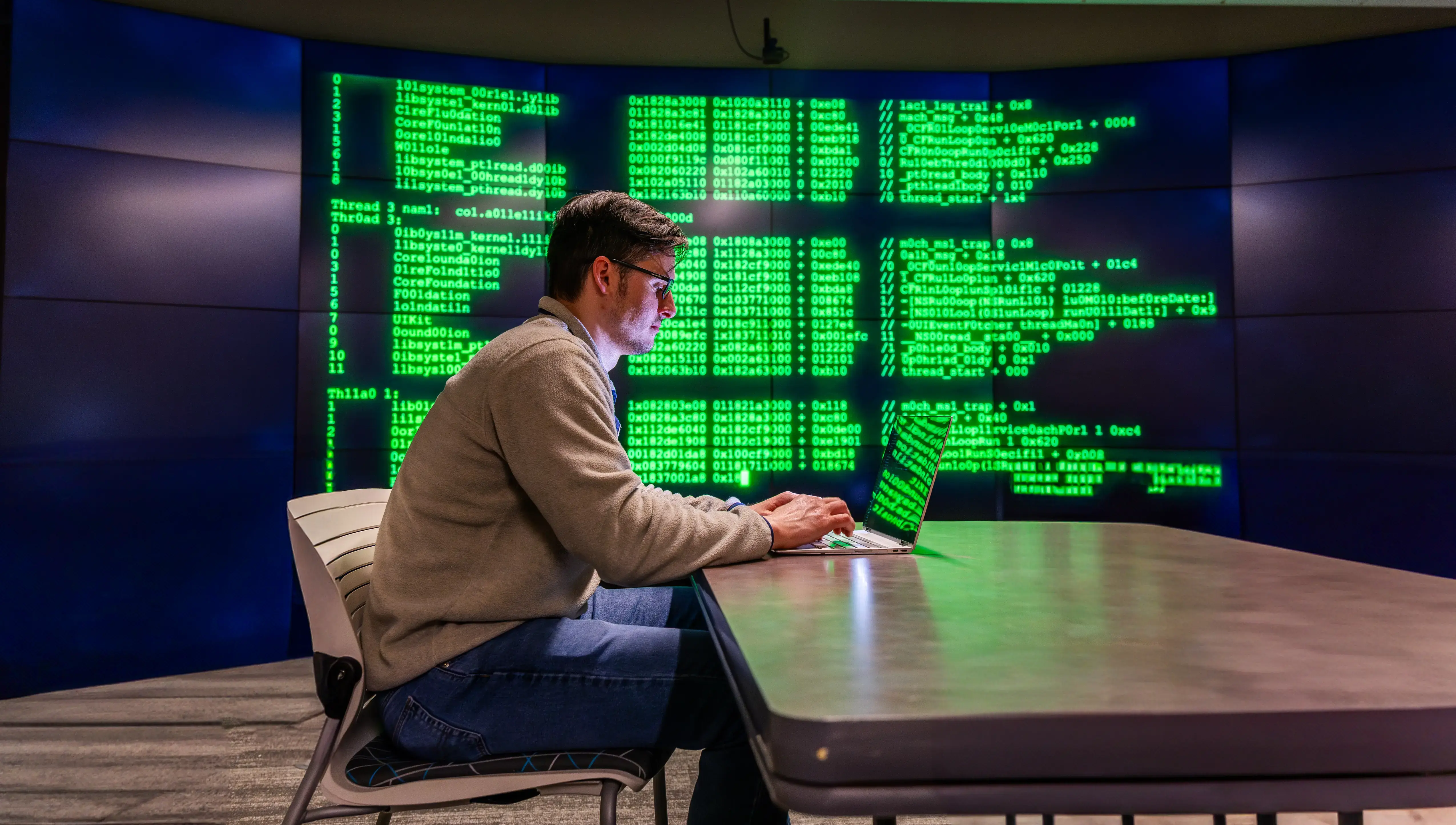 A man types on a laptop in a dim room as green code reflects on his screen and a large wall display behind him.
