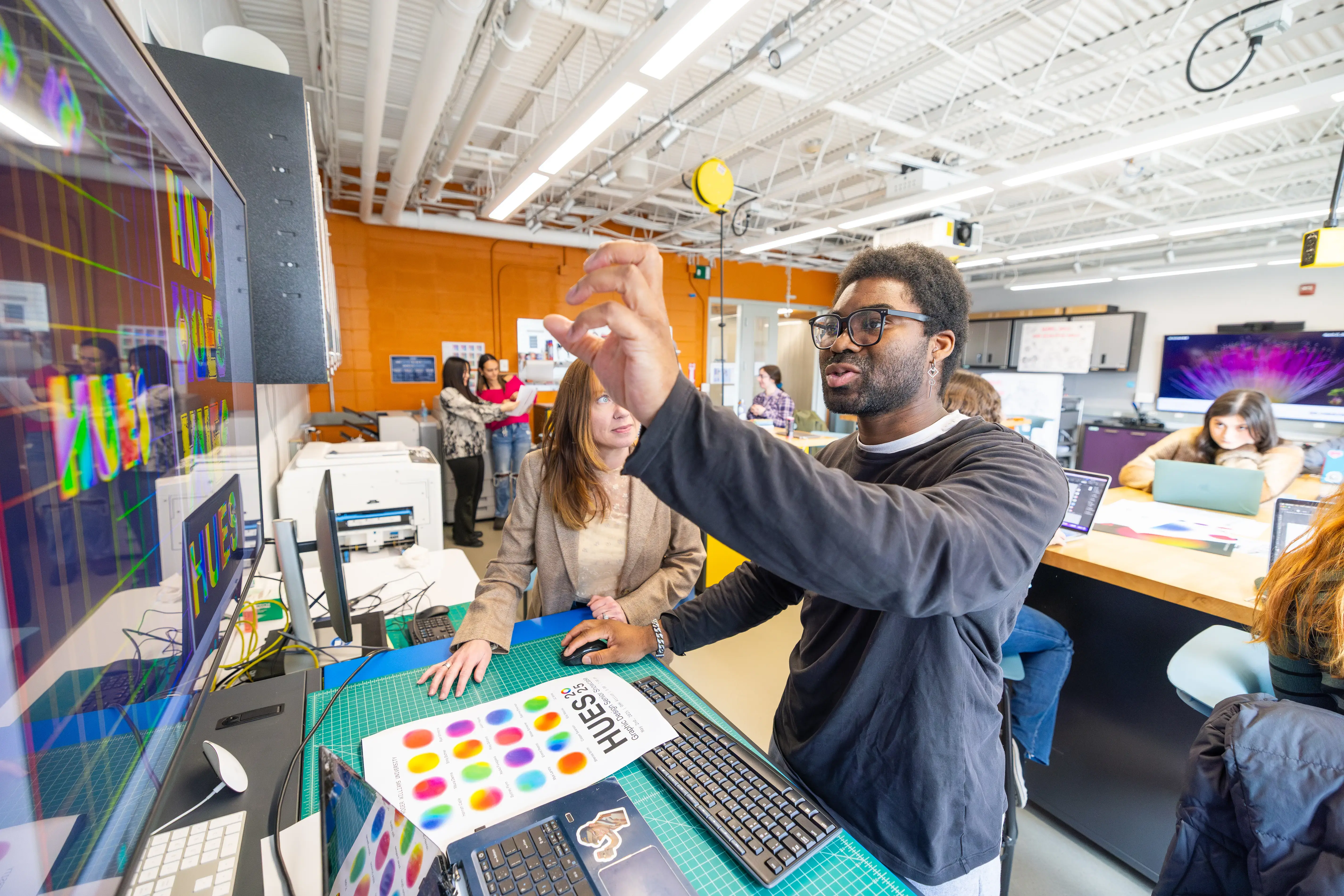 Students work on graphic design in a modern classroom as a man gestures to a monitor and speaks with a woman beside him.