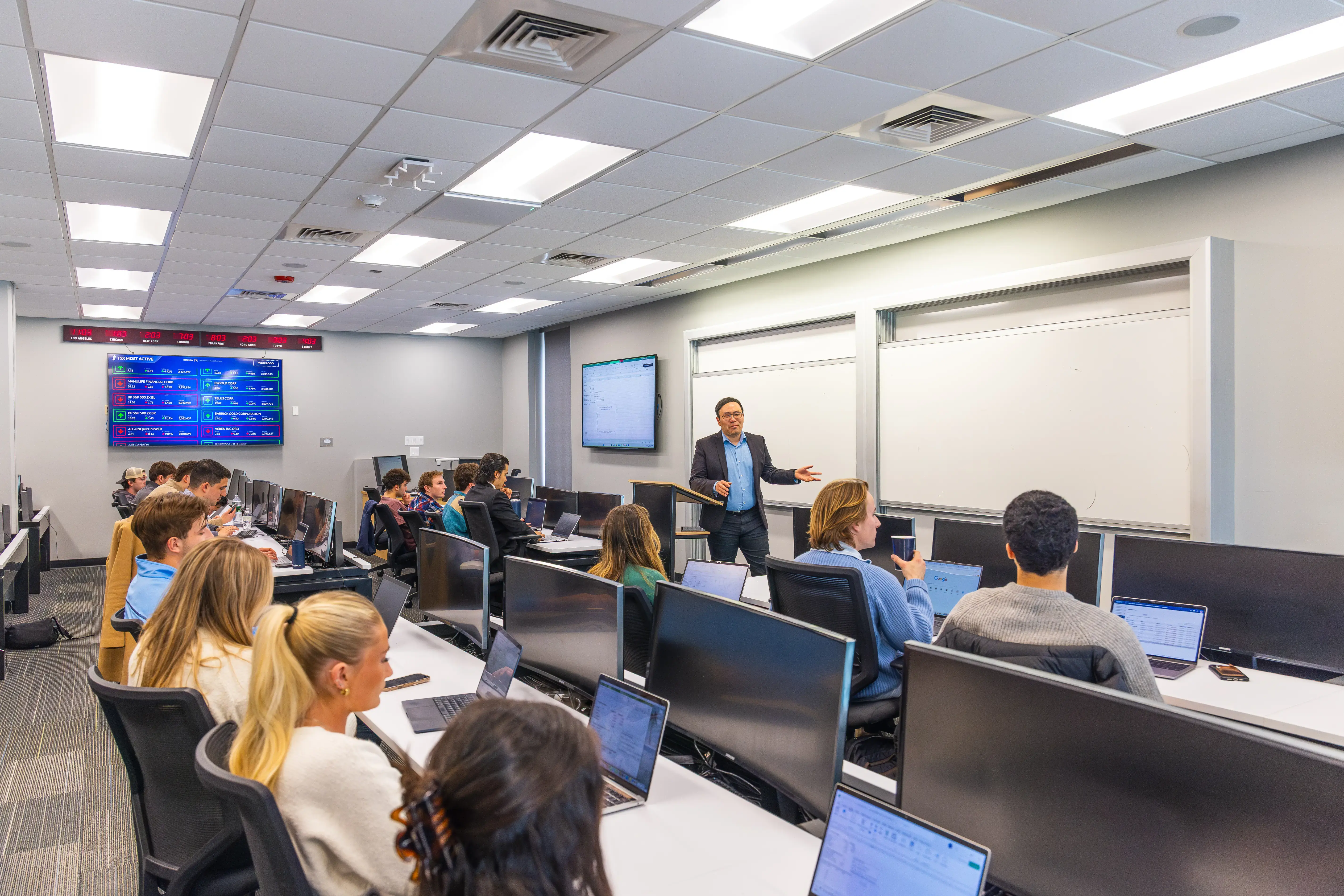 College classroom with students seated at desks, laptops open, listening to a professor speaking at the front.