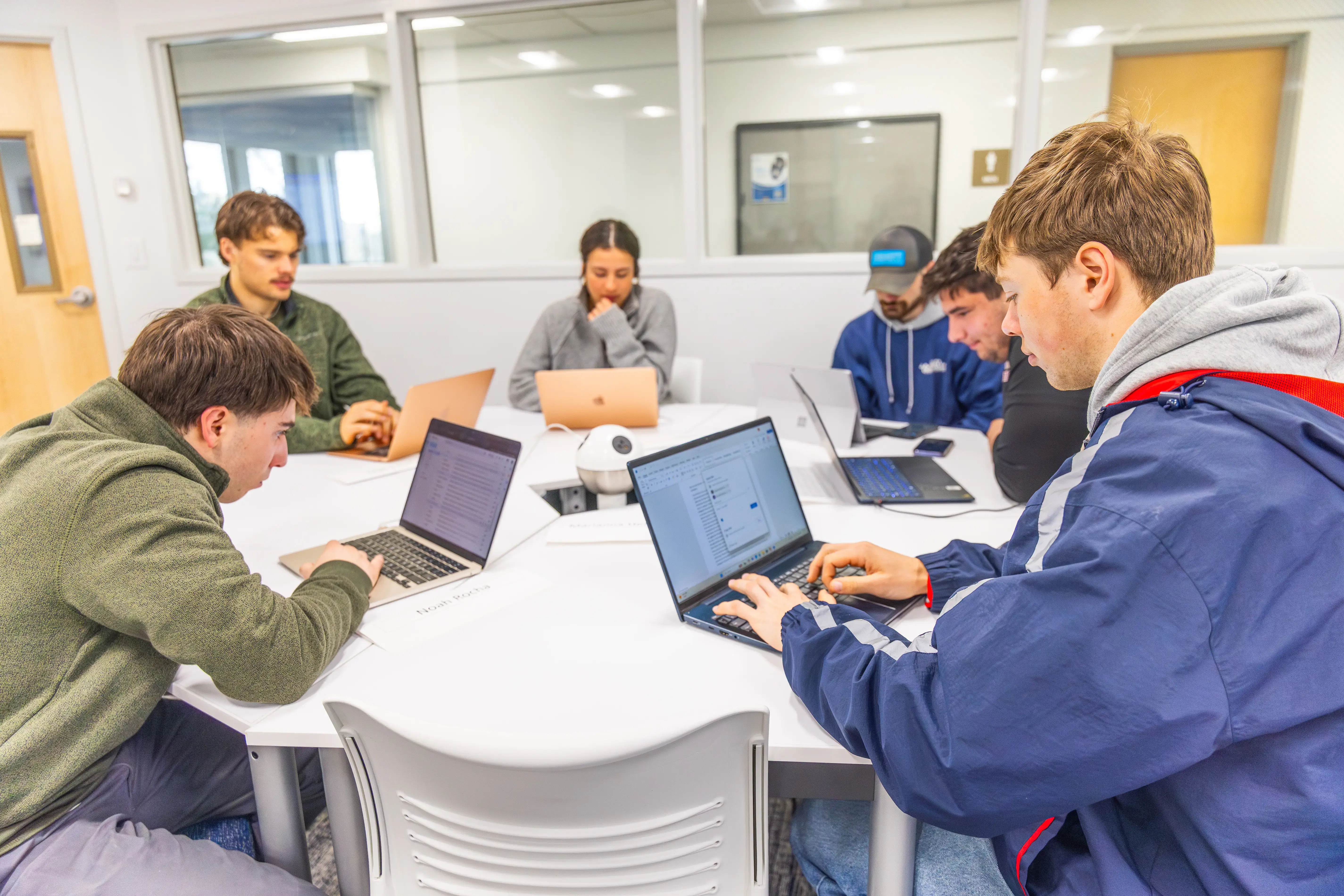 Group of college students in casual clothes working on laptops around a white table in a bright study room.