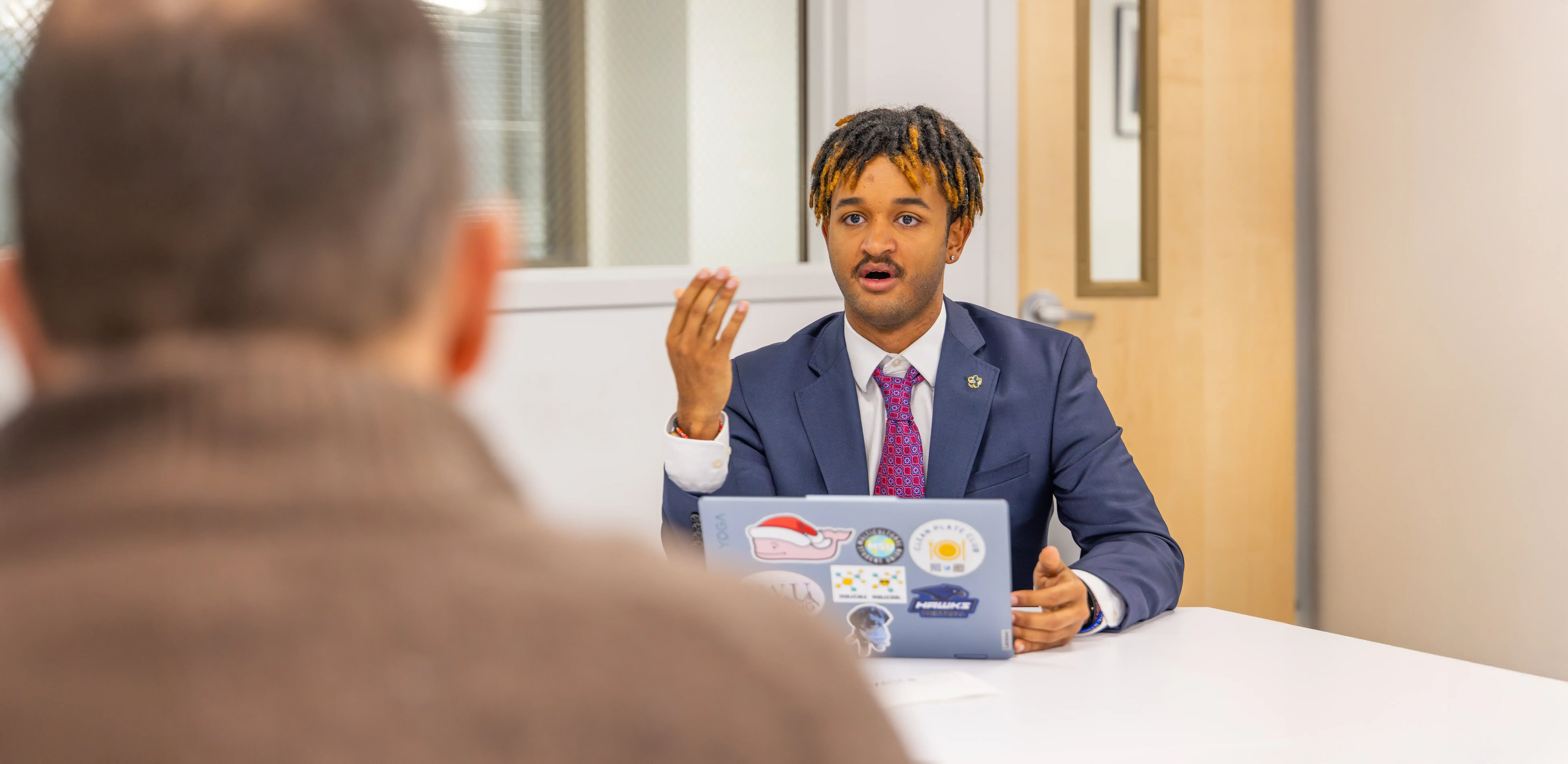 A student in a suit gestures while speaking during a meeting or interview, with a sticker-covered laptop open on the table.