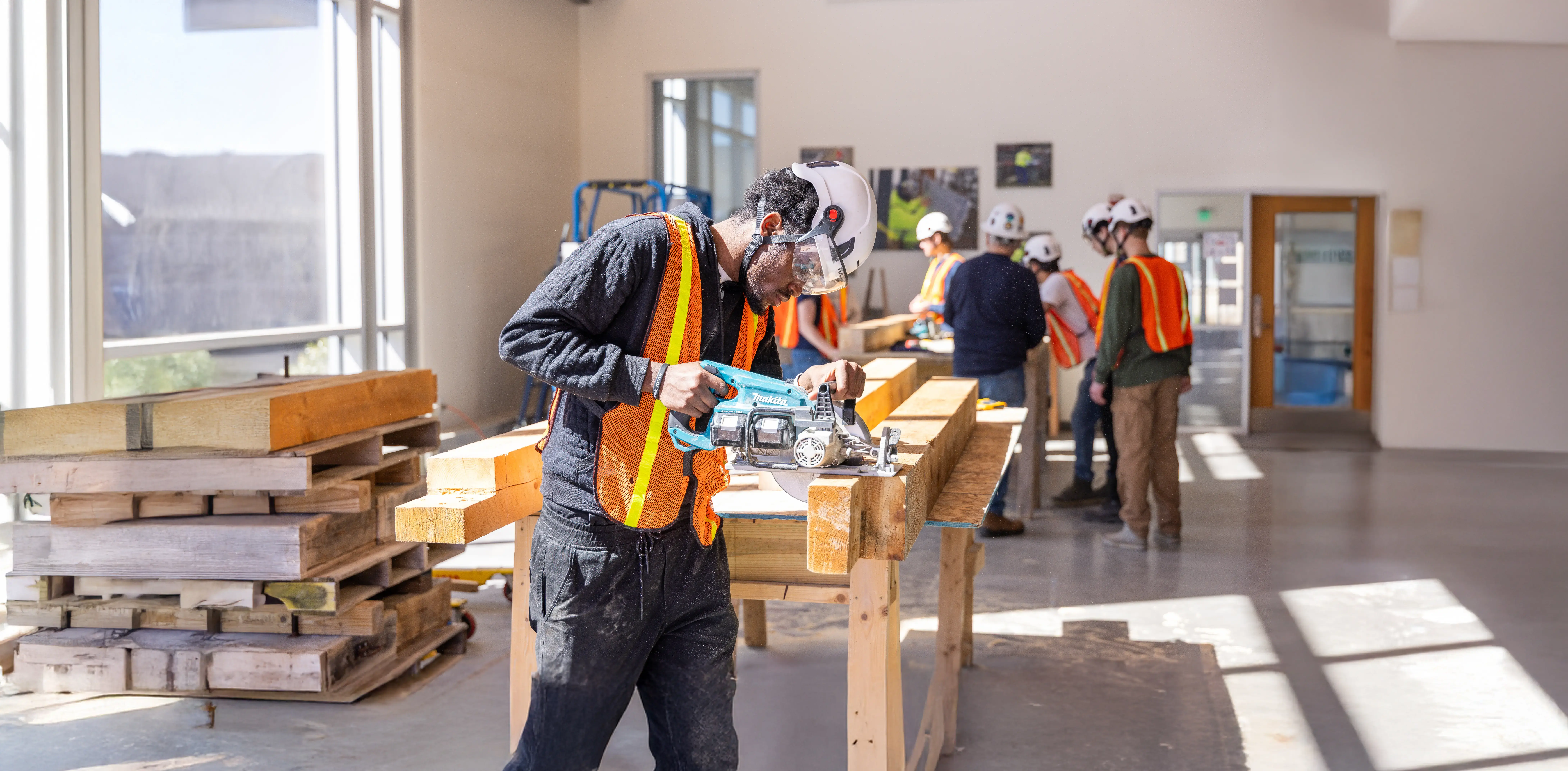 Man wearing safety gear cuts wood with power saw in a bright workshop while others work on carpentry projects nearby.
