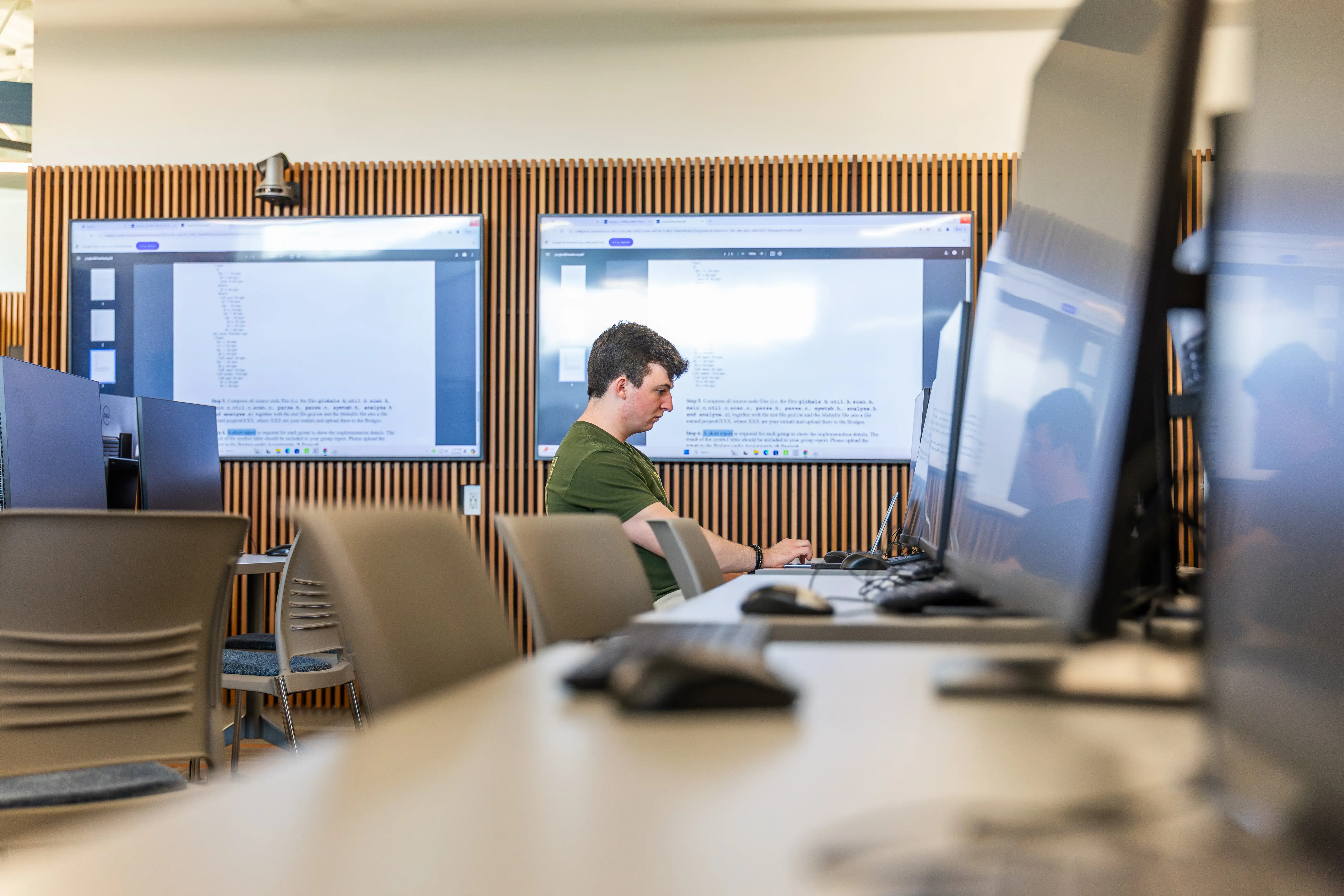 A young man works on a laptop in a modern lab with code on screens behind him and monitors in the foreground.