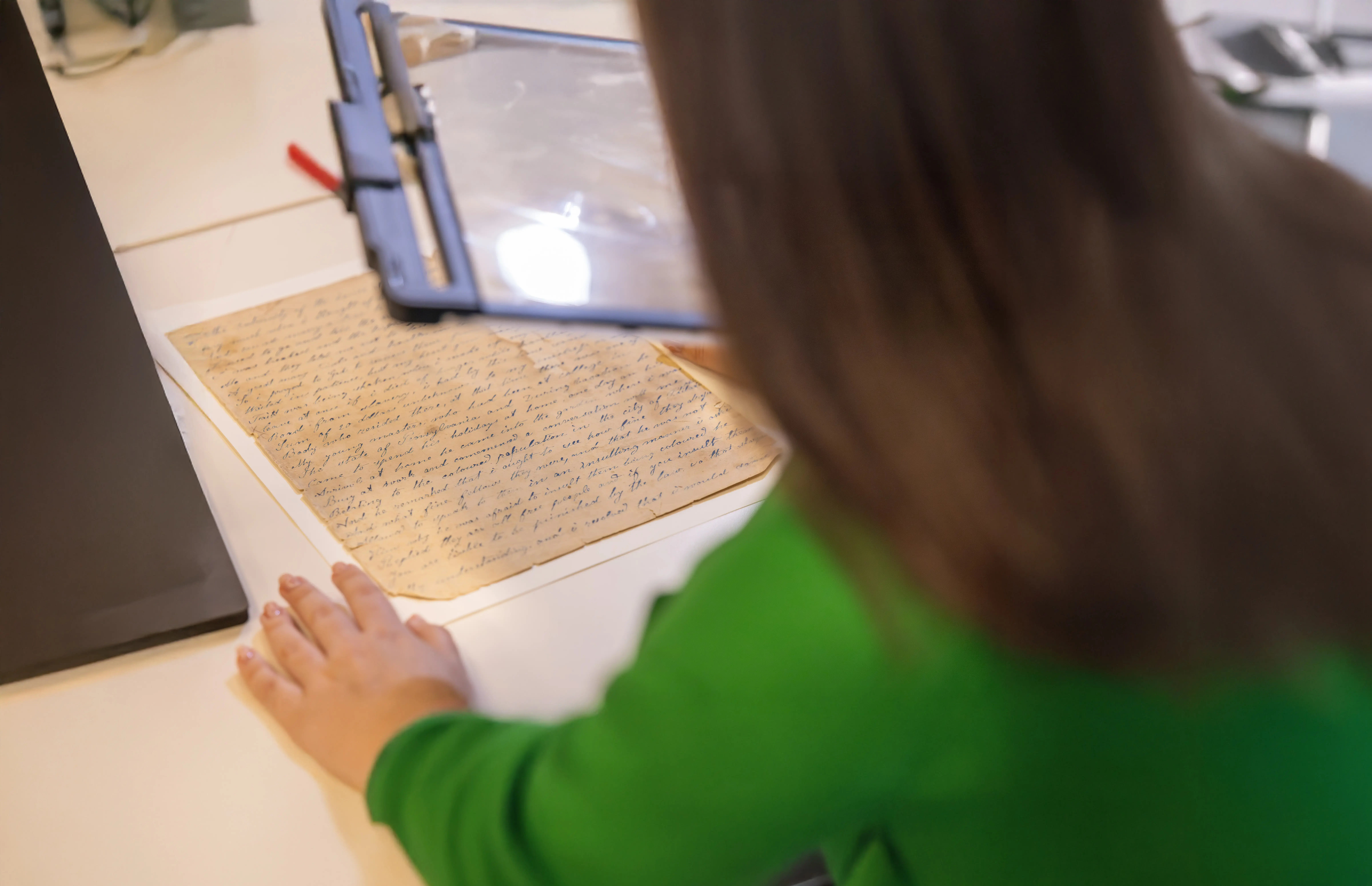 A person with a green sleeve uses a magnifying glass to examine an old handwritten document on yellowed paper.