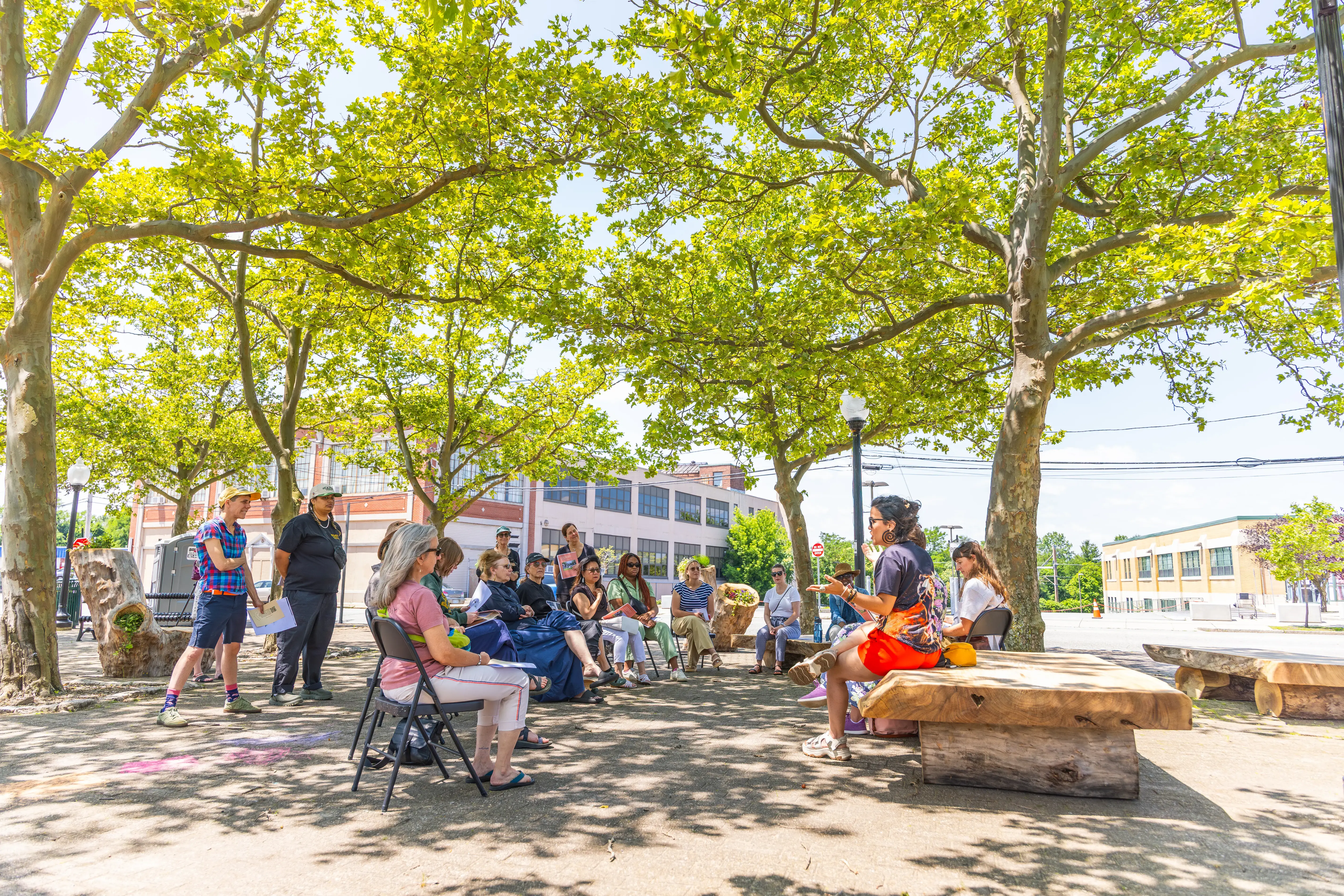 A group sits outdoors in a shaded circle under trees, engaged in a discussion on a sunny day near urban buildings.