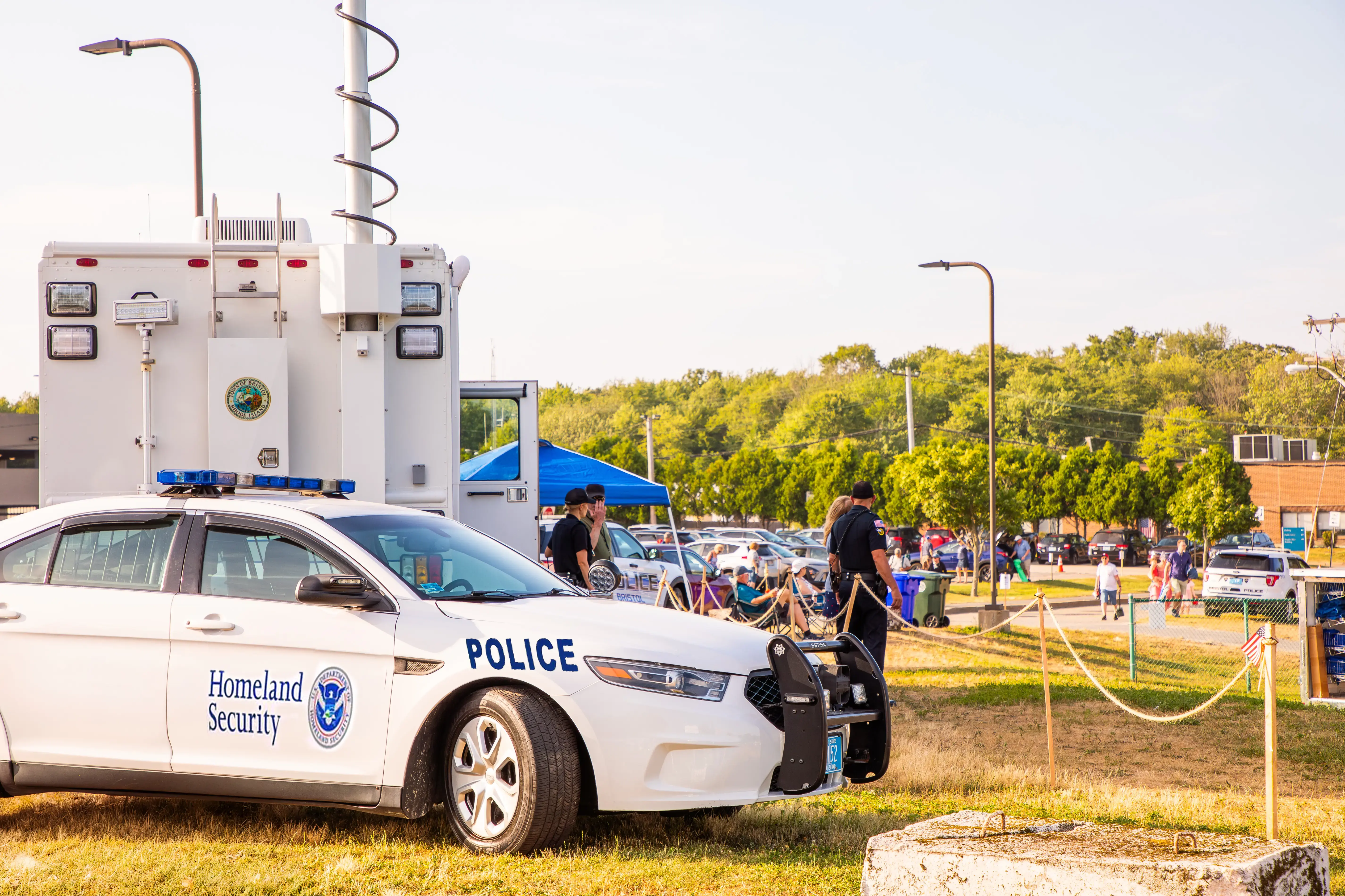 A Homeland Security police car and mobile unit are parked at a public outdoor event with people nearby.