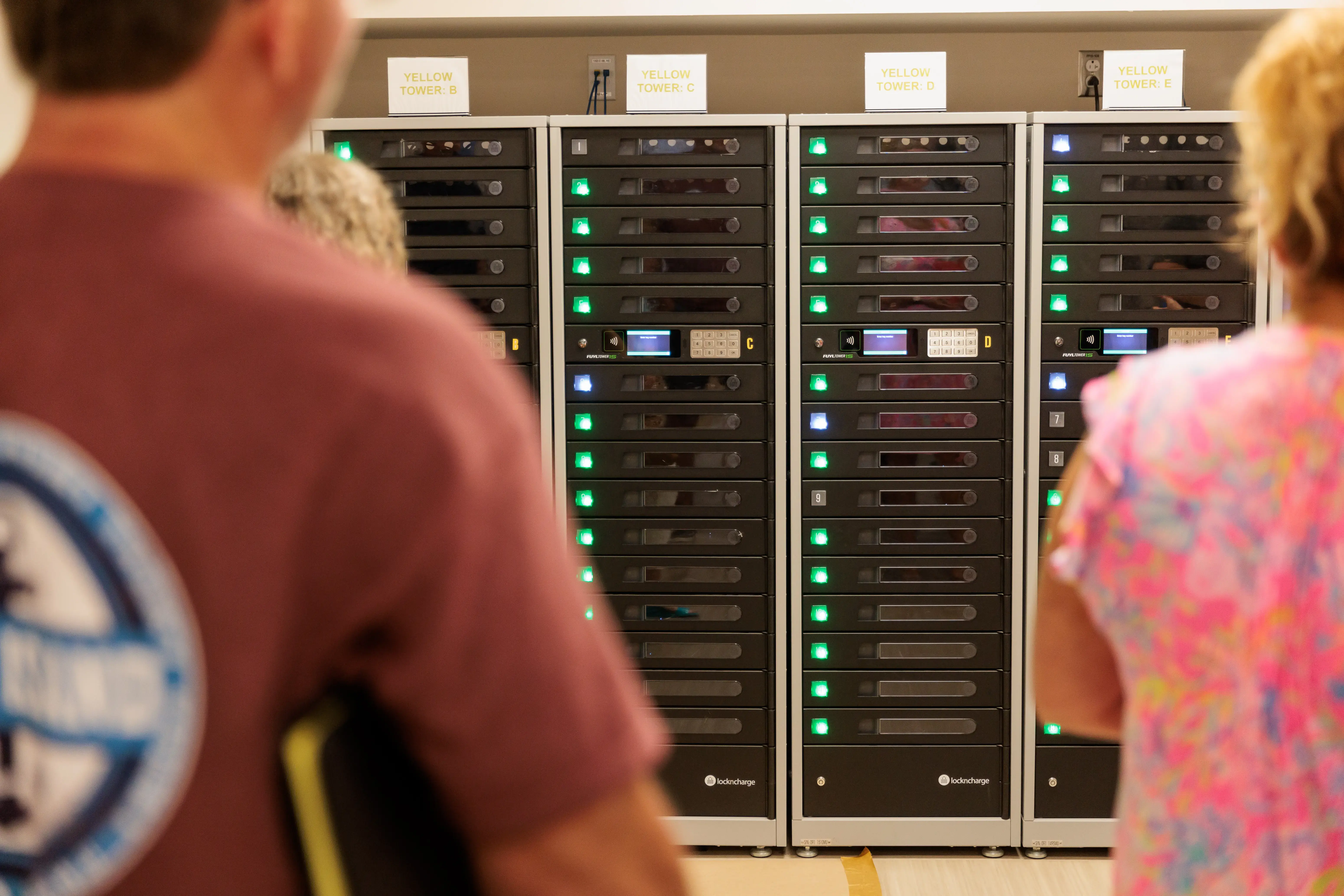 People stand by secure charging lockers labeled “Yellow Tower” with compartments featuring lights, keypads, and screens.