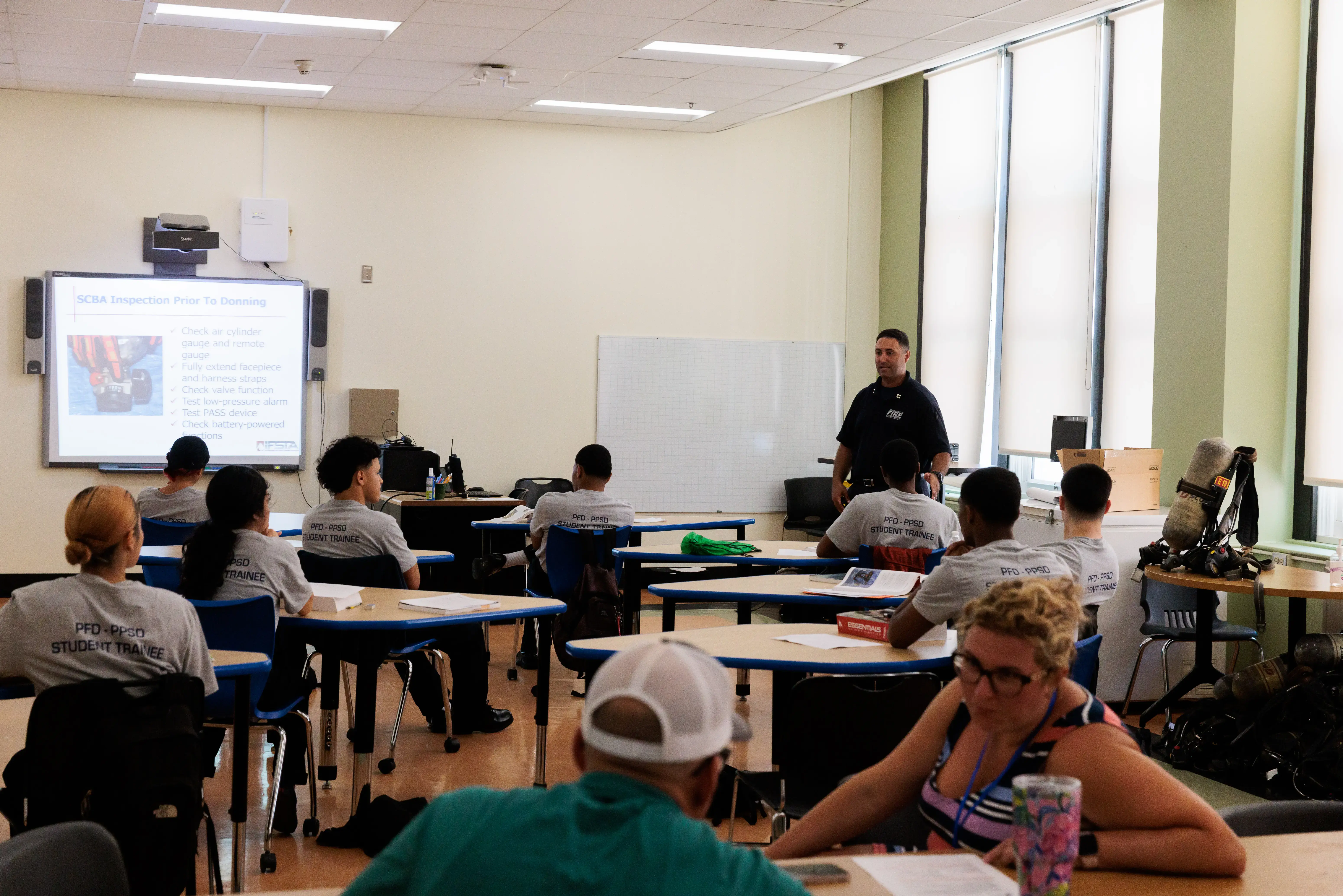 Student trainees in gray shirts listen to an instructor teaching n in a fire training classroom with equipment nearby.