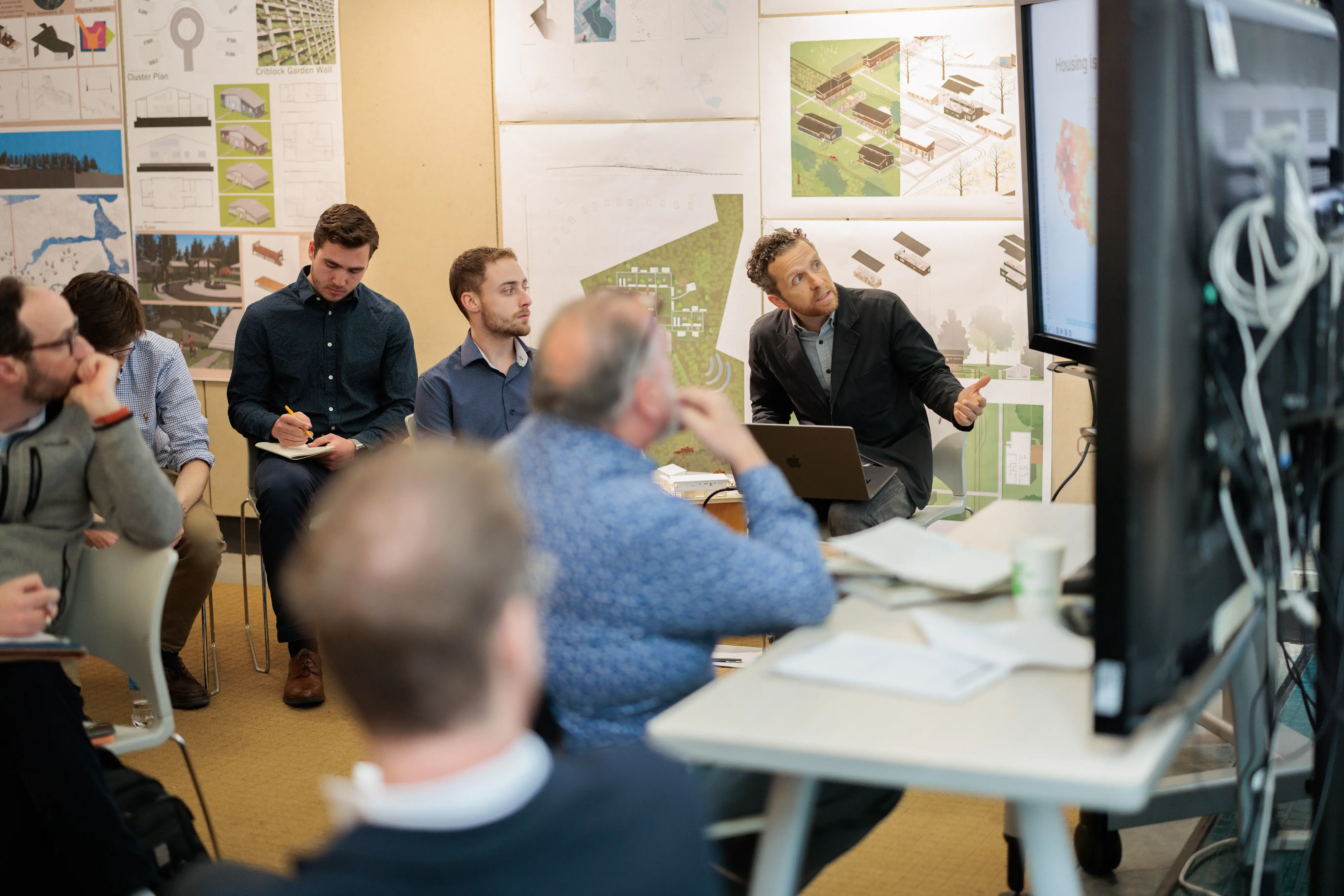 People in a design studio listen as a man presents architectural plans on a screen amid colorful drawings on the walls.