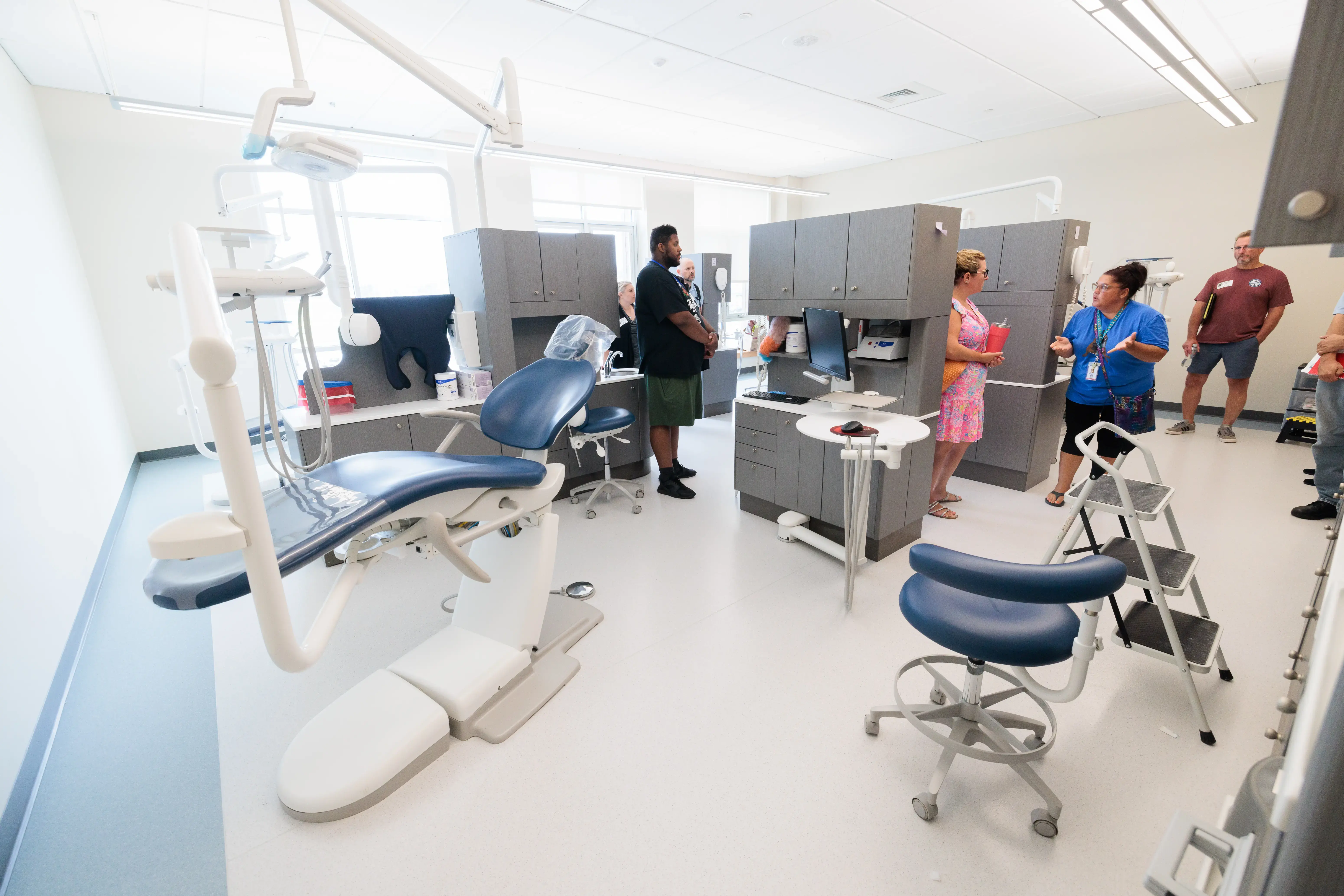 A woman in a blue shirt explains a modern dental clinic room to visitors, with chairs, tools, and white walls and floors.