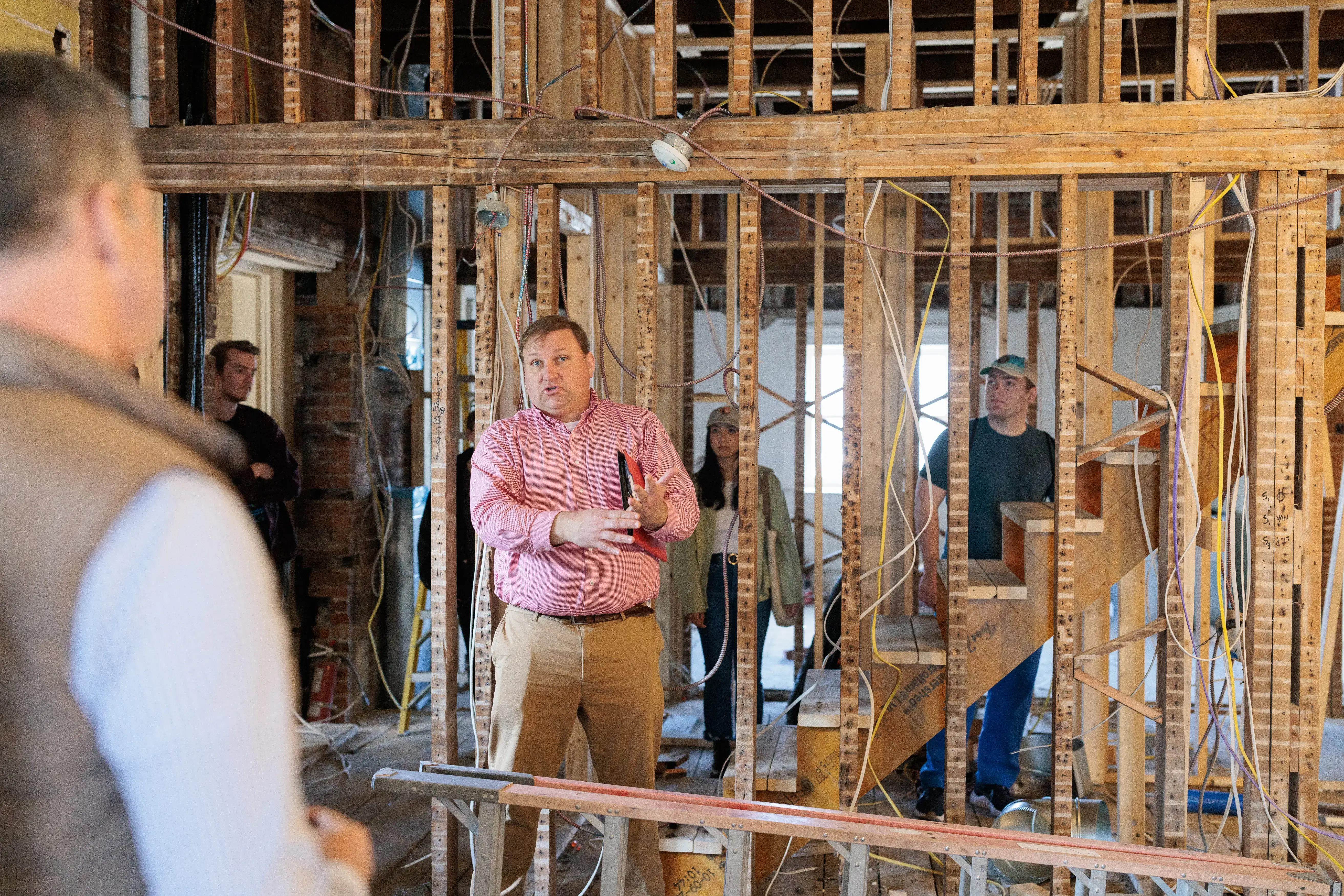A man speaks to a group inside a building under construction, surrounded by exposed wood framing and unfinished stairs.