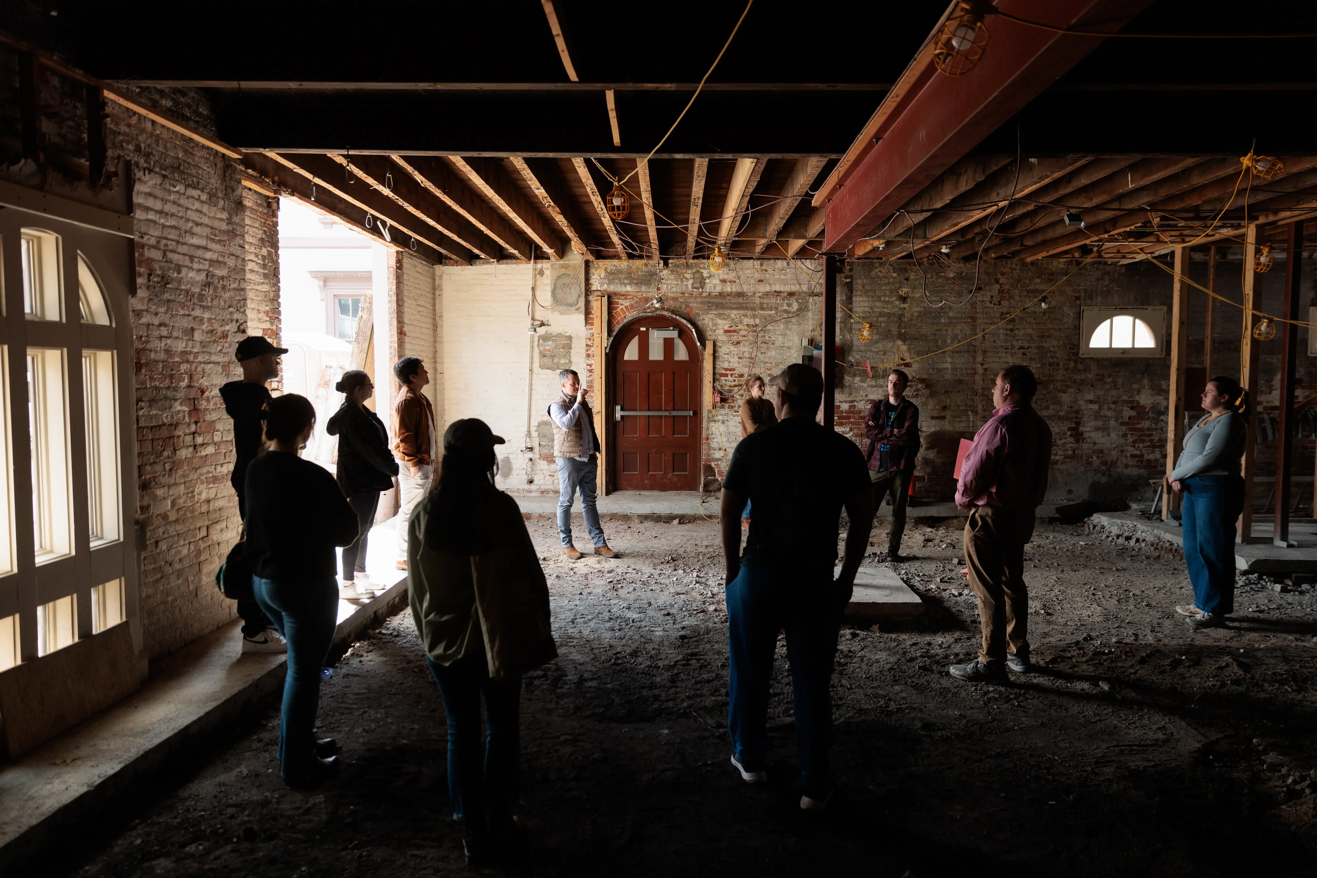 A group stands in a dimly lit, partially renovated brick building as one person speaks, giving a construction site tour.