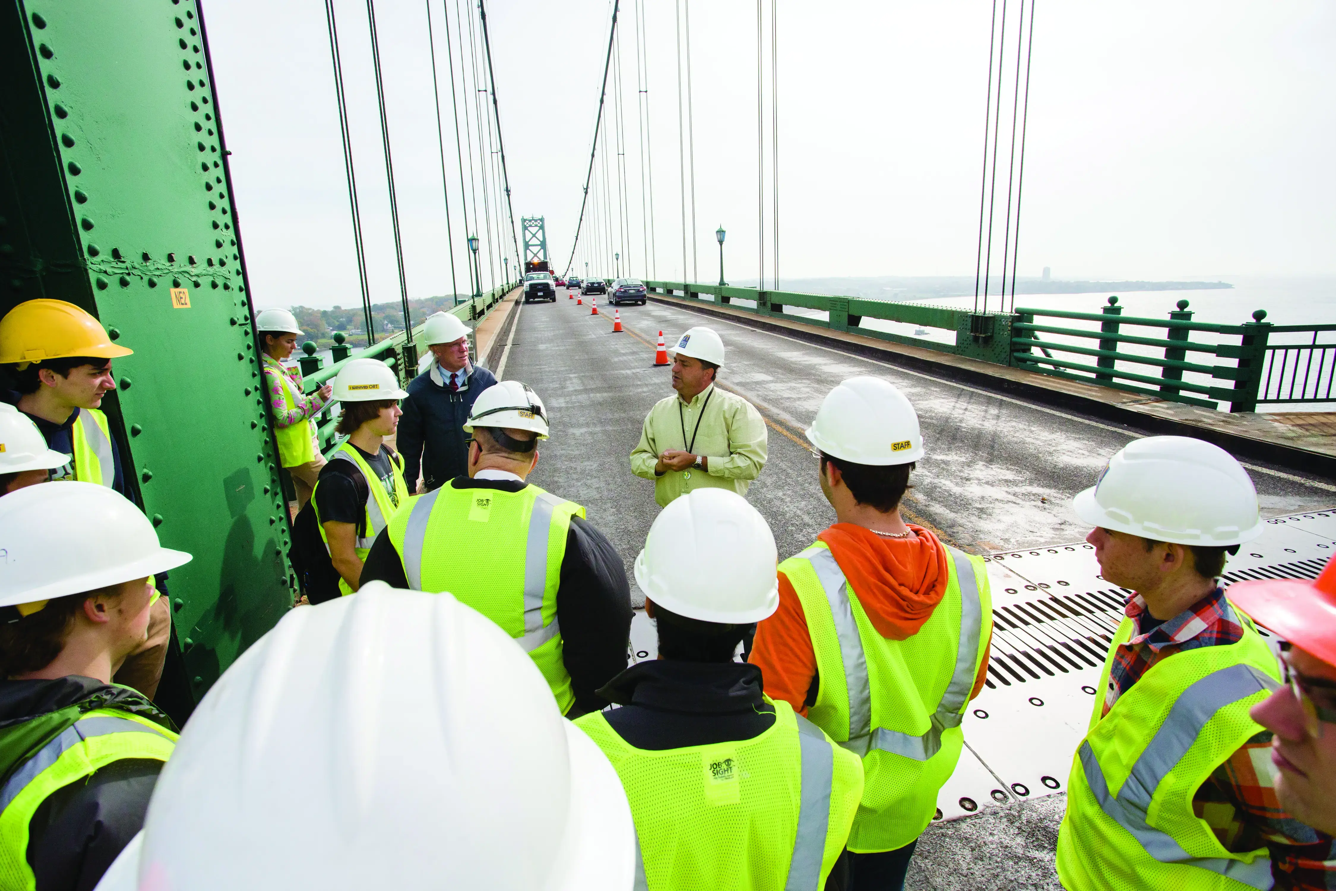 A group of civil engineering students in hard hats and vests gathered on a bridge, listening to a professor.