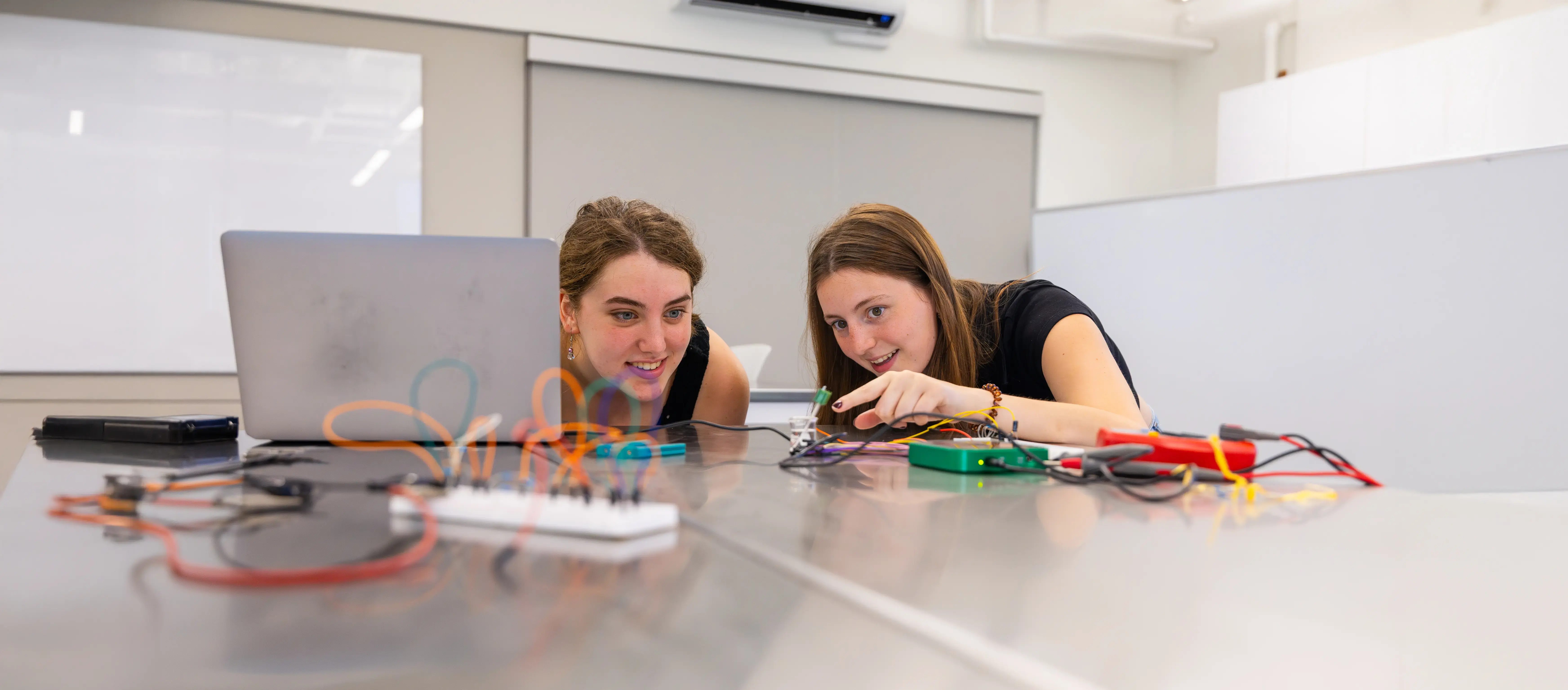 Two students work together on an electronics project, smiling as they test circuits with wires on a table beside a laptop.