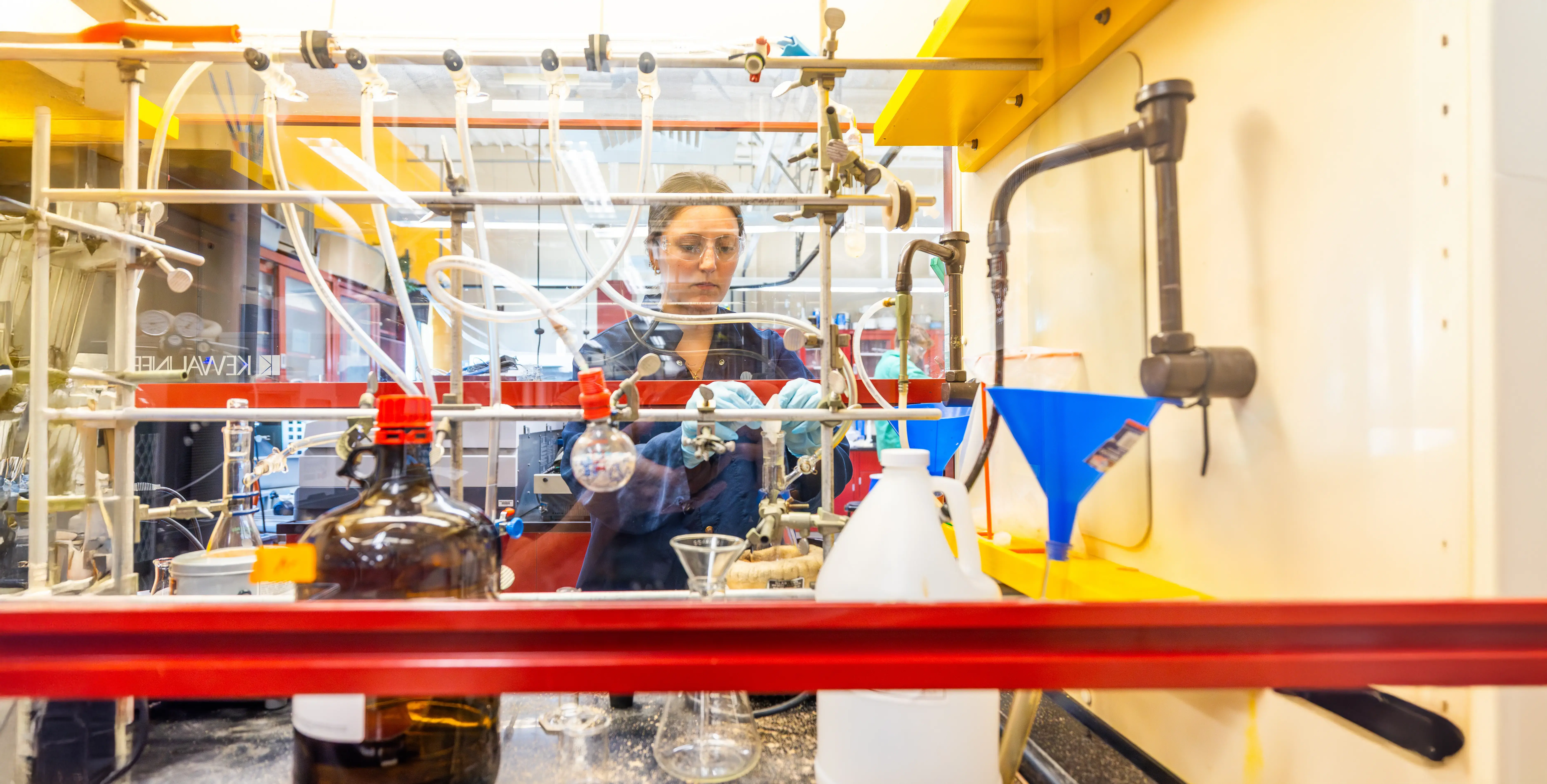 A student in safety gear works behind a fume hood with tubes, flasks, and lab equipment.