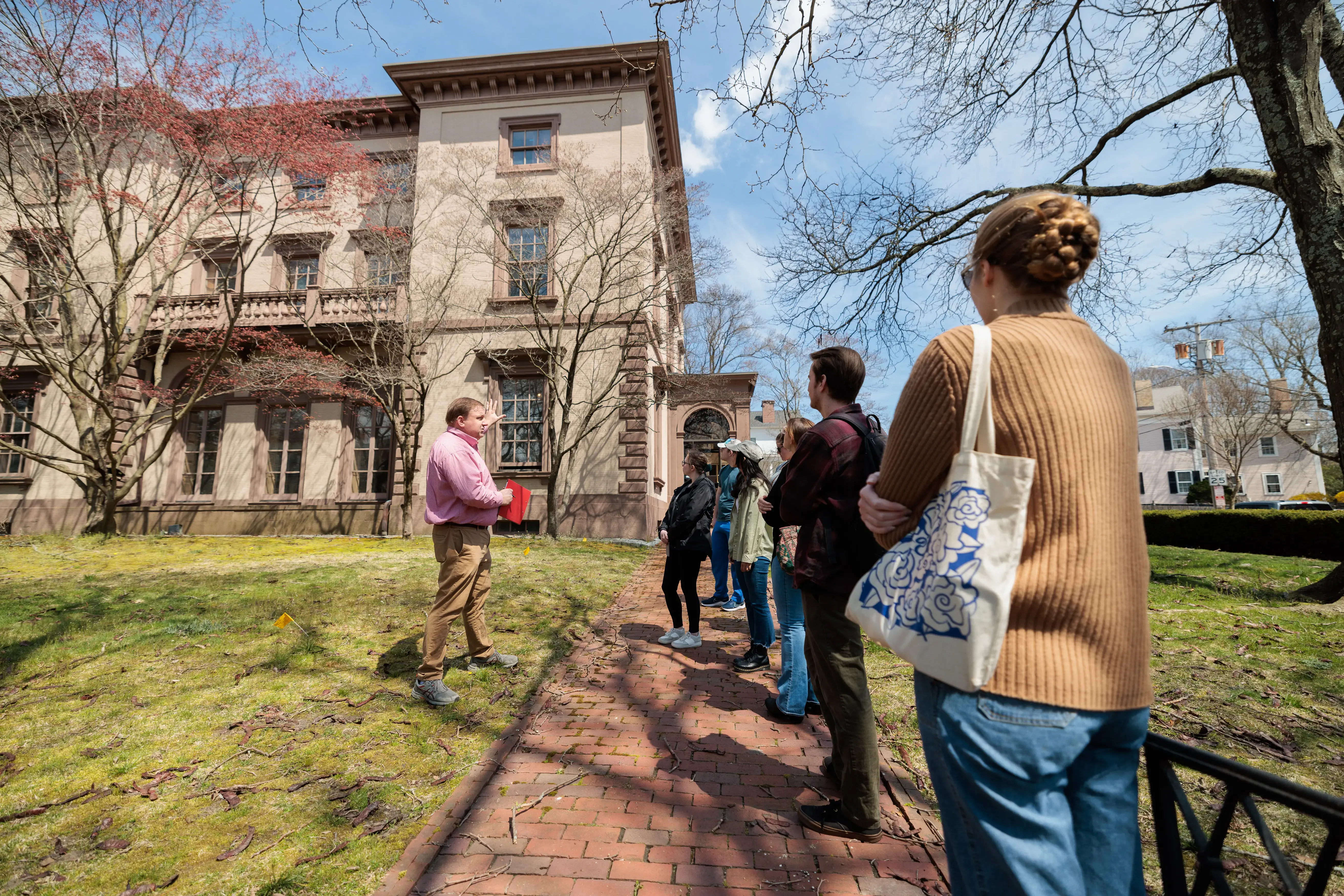 Tour guide speaks to a group outside a historic building on a sunny day, with trees and brick path in view.