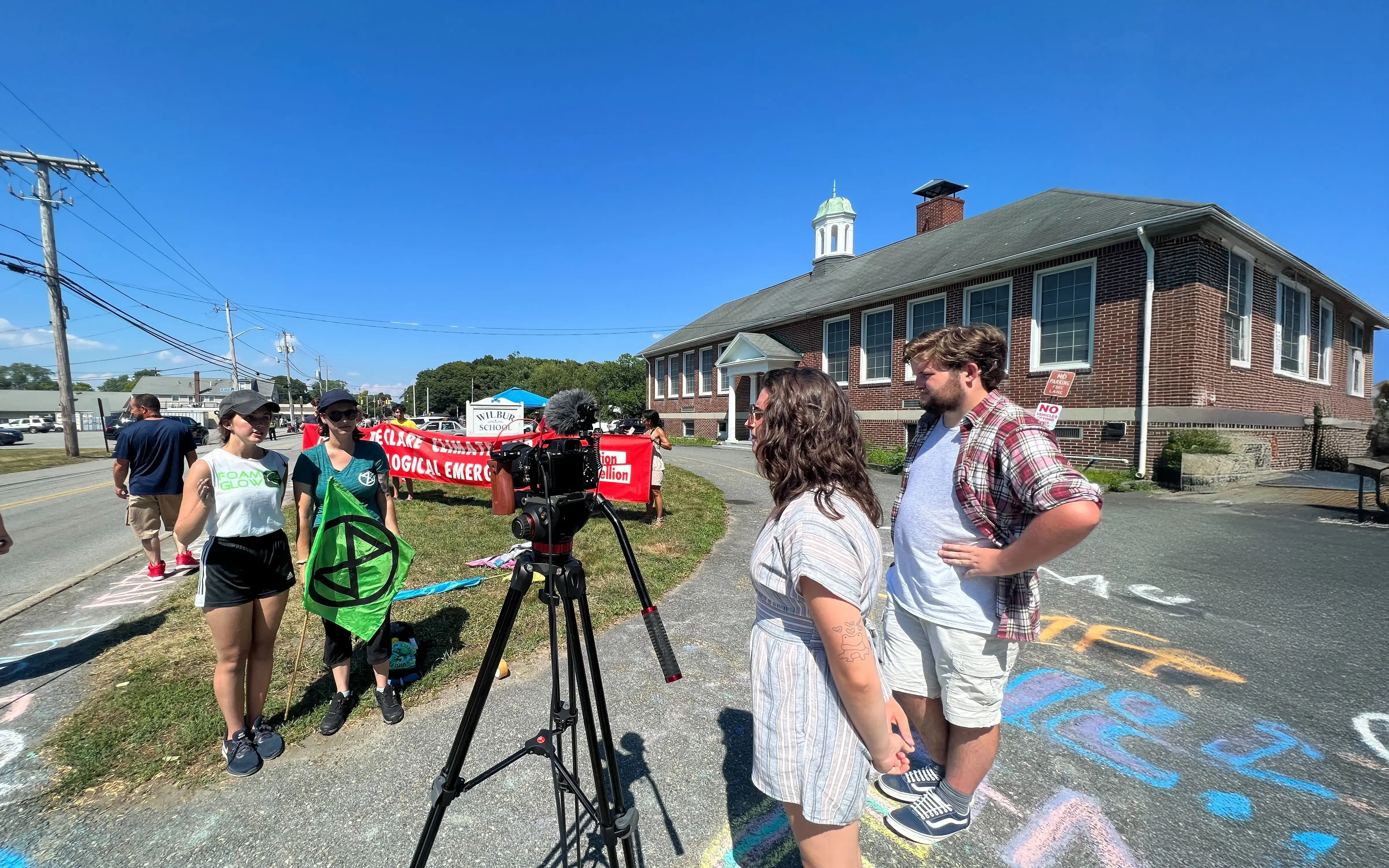 People record an environmental protest outside a brick building, with signs, banners, and chalk art on the ground.