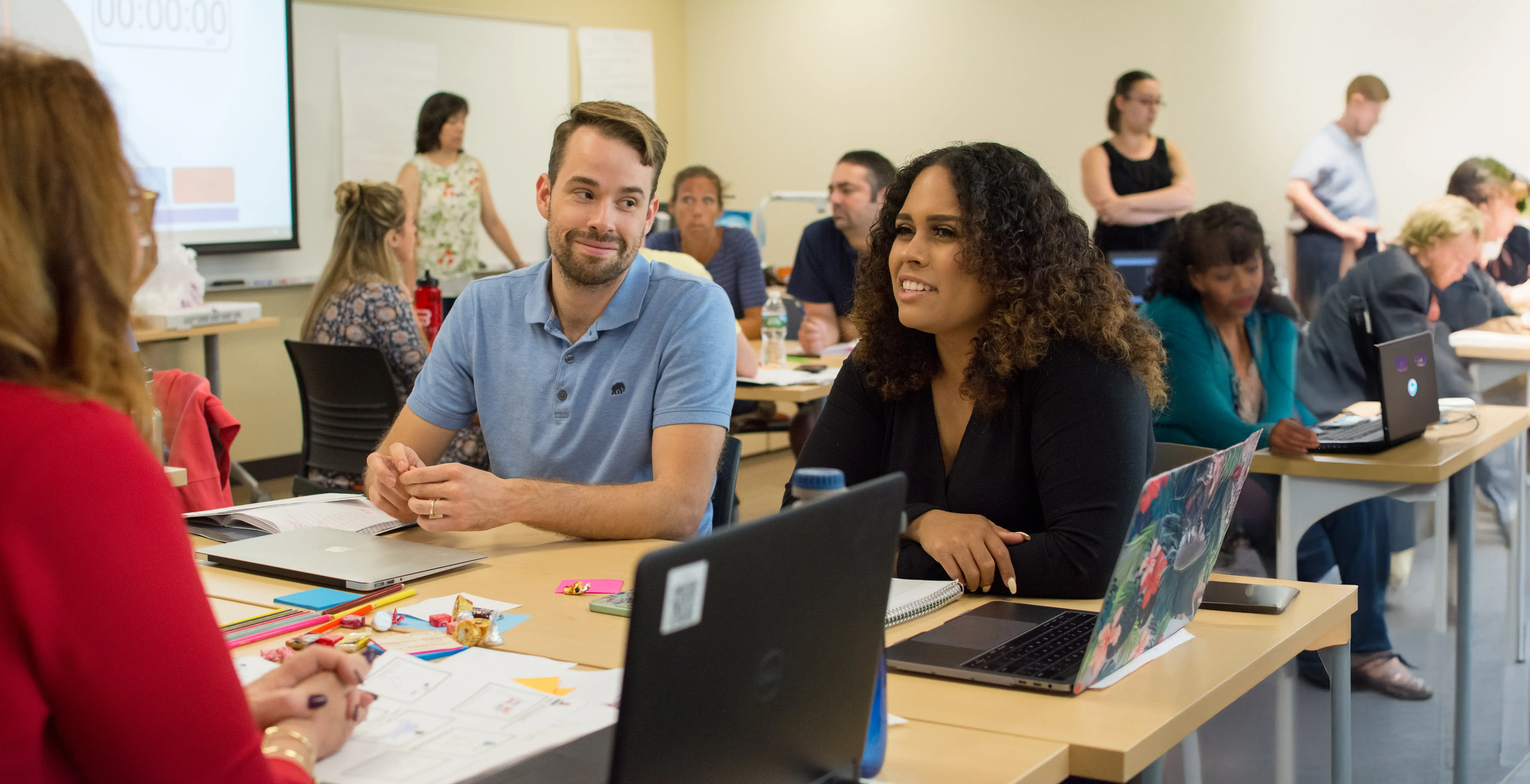 ESL educators collaborate in a classroom, engaging in group work with laptops, notes, and a timer projected on the board.