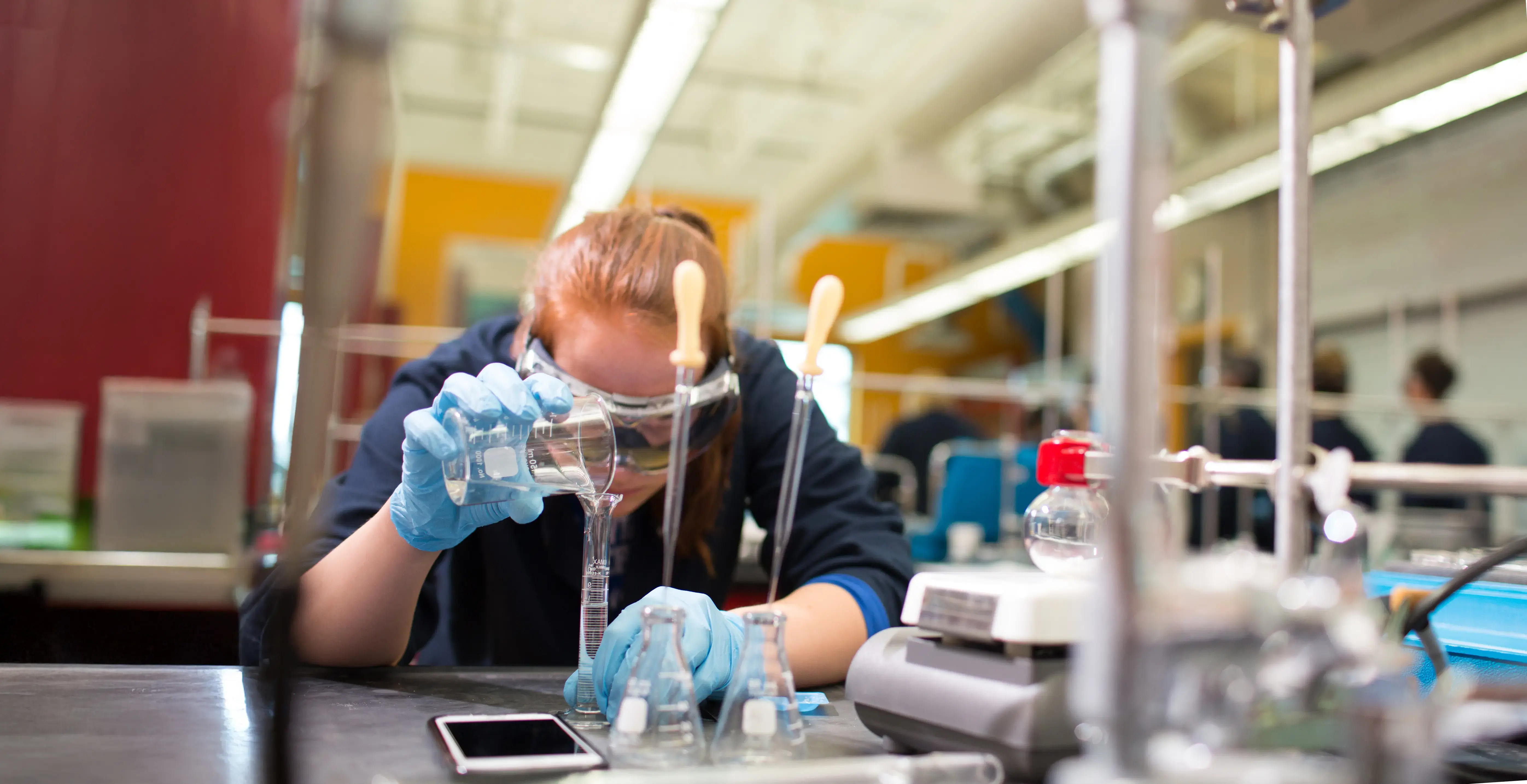 A student in lab coat and gloves pours liquid into a graduated cylinder in a science lab.