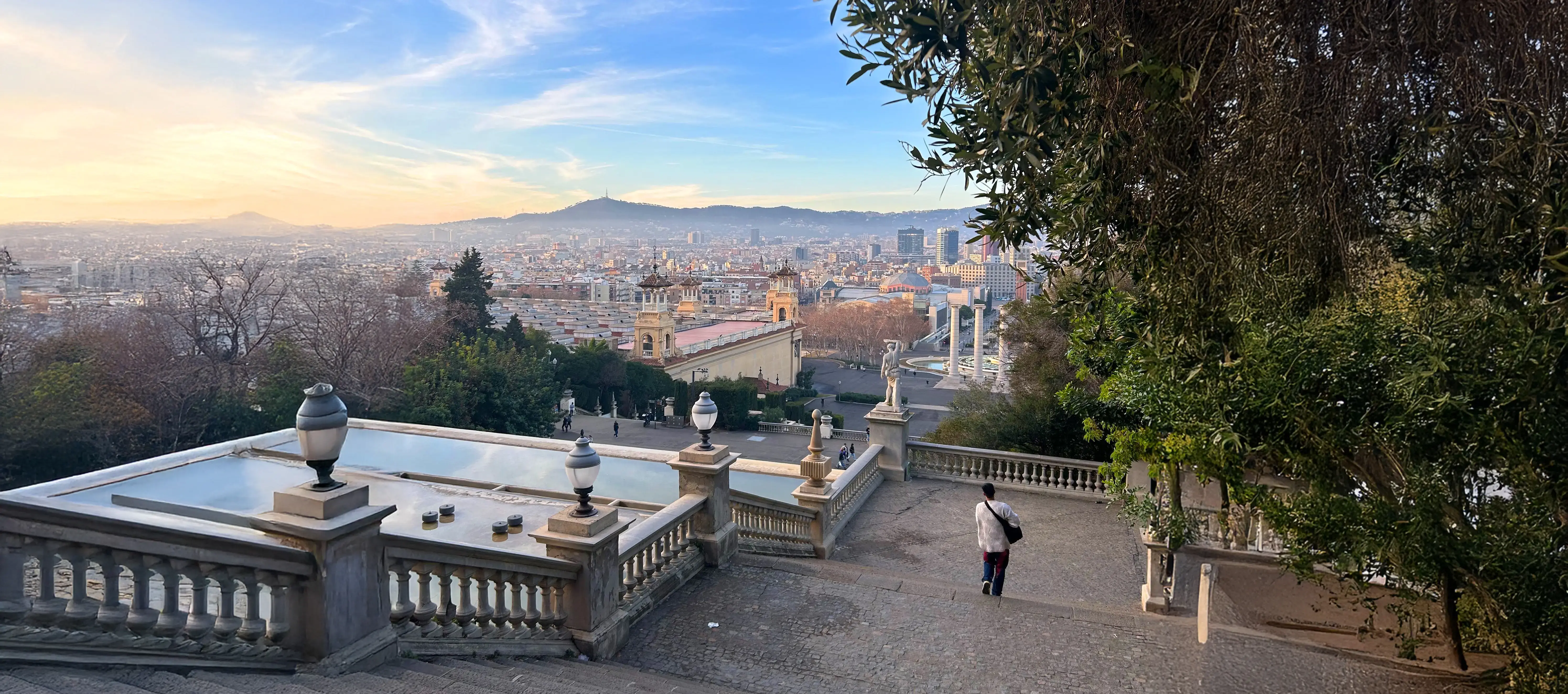 A person walks down terrace steps overlooking Barcelona, Spain at sunset, with cityscape, trees, and mountains visible.