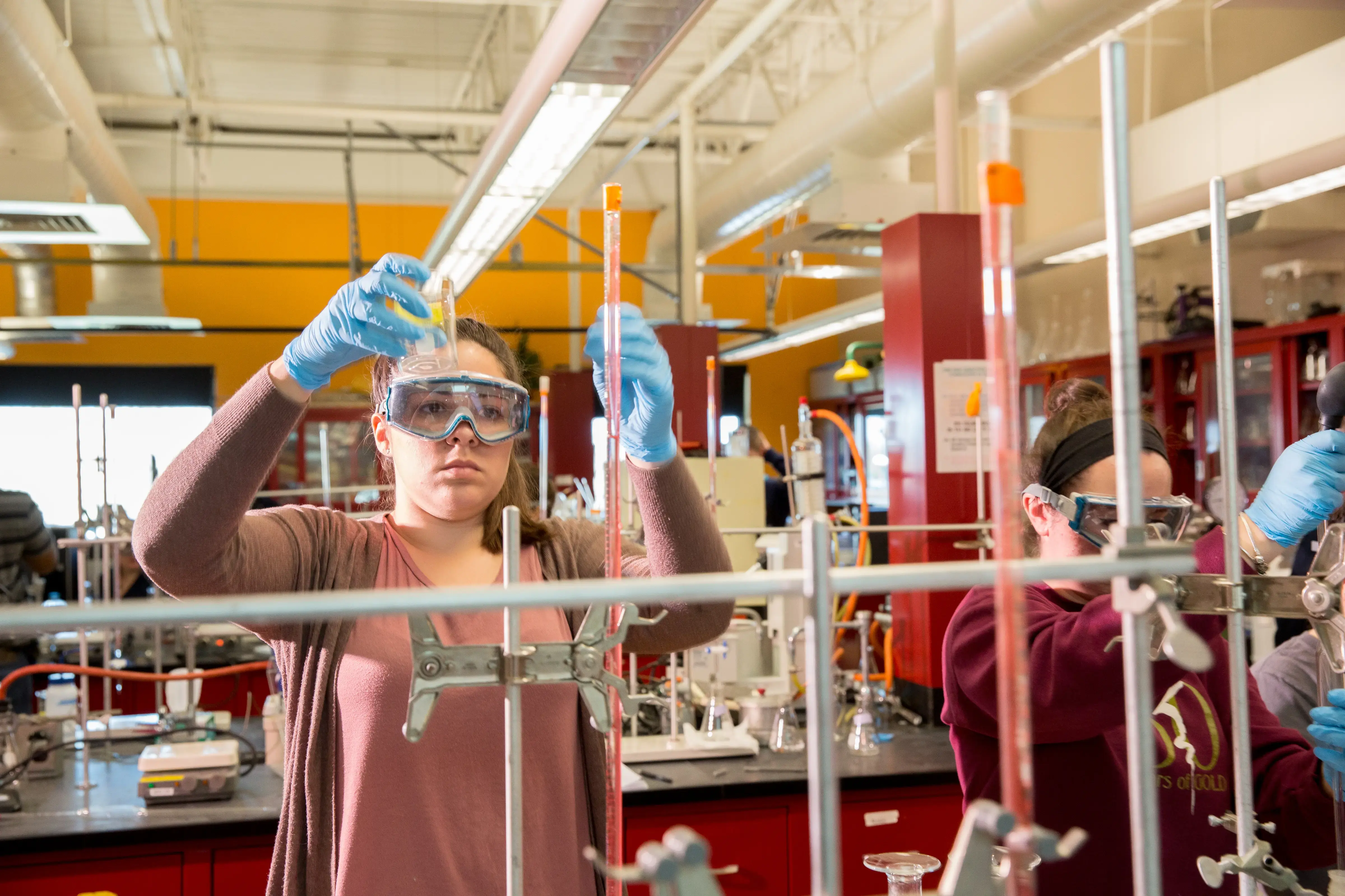 Two students in goggles and gloves conduct a chemistry experiment in a bright lab with beakers, tools, and red cabinets.